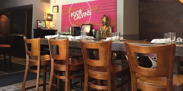 Un comedor con sillas de madera y una mesa lista para una comida. Se ve una estatua de Buda y un letrero que dice “Poor Calvin's” al fondo.