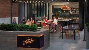 An outdoor seating area with tables and chairs, a bar with shelves of bottles, and a sign that reads "El Vinedo Local".