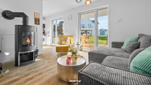 A modern living room with a wood-burning stove, a round coffee table with decorative items, a grey sofa, and a view of the outdoors through large windows.