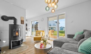 Un salon moderne avec un poêle à bois, une table basse ronde agrémentée d’objets décoratifs, un canapé gris et une vue sur l’extérieur grâce à de grandes fenêtres.