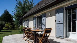 Une terrasse extérieure avec une table et des chaises, un sol carrelé et un bâtiment doté de volets et d’une fenêtre.
