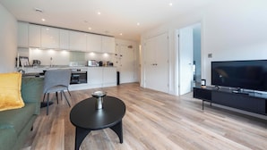 A modern living room with a sofa, a coffee table, a TV, and a kitchen area with cabinets and appliances. - Modern 1 Bed Apartment in Gravesend w Games Room (Gravesend)