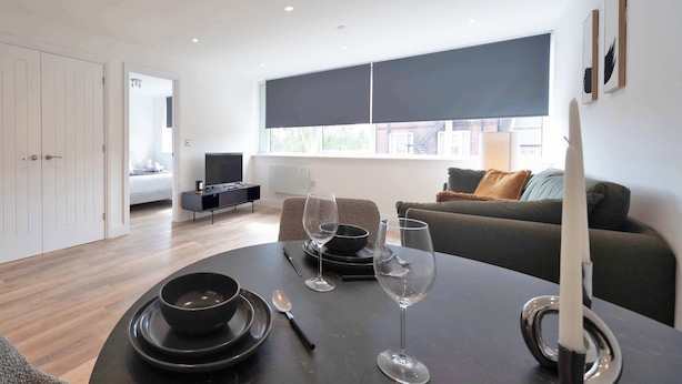 A modern living room with a dining table set for two, a sofa, and a television in the background. - Modern 1 Bed Apartment in Gravesend w Games Room (Gravesend)