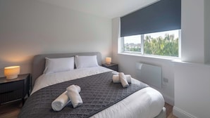 A modern bedroom with a large bed, a window with a view, and a bedside table with a lamp. - Modern 1 Bed Apartment in Gravesend w Games Room (Gravesend)
