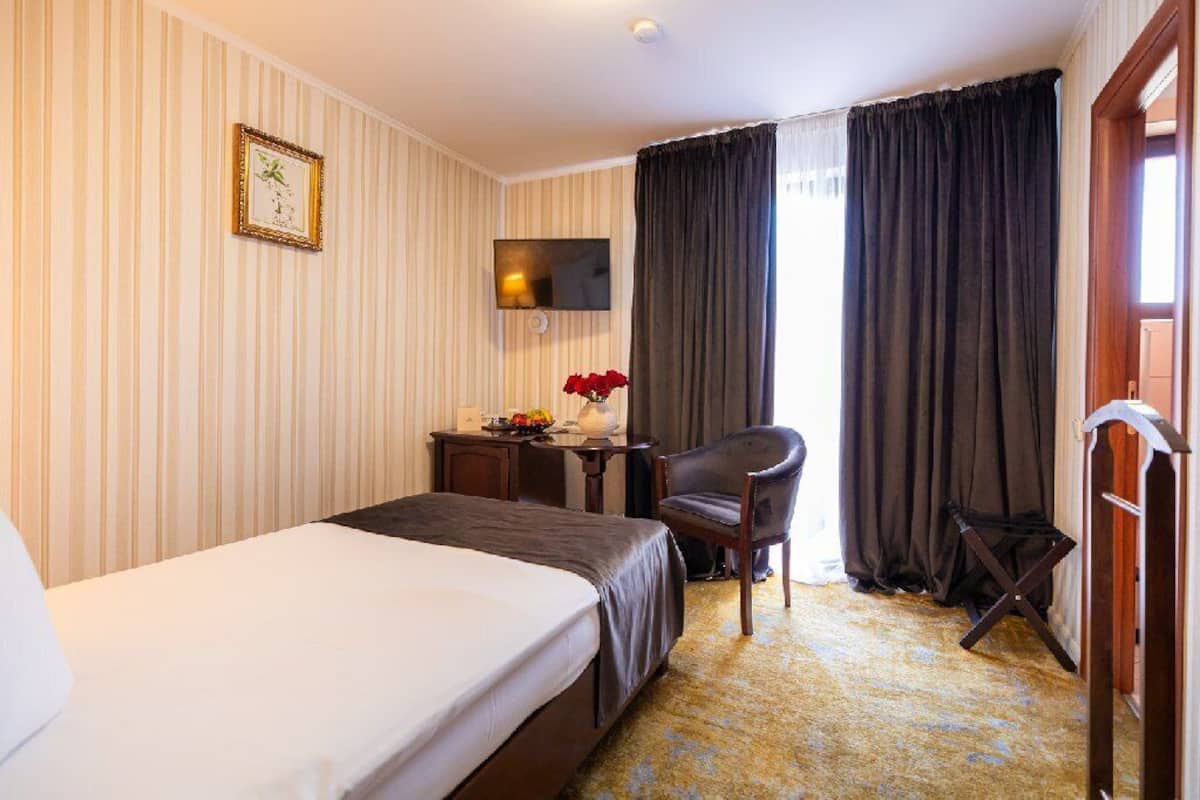 A hotel room with a bed, a desk with a chair, a TV mounted on the wall, and a window with curtains.