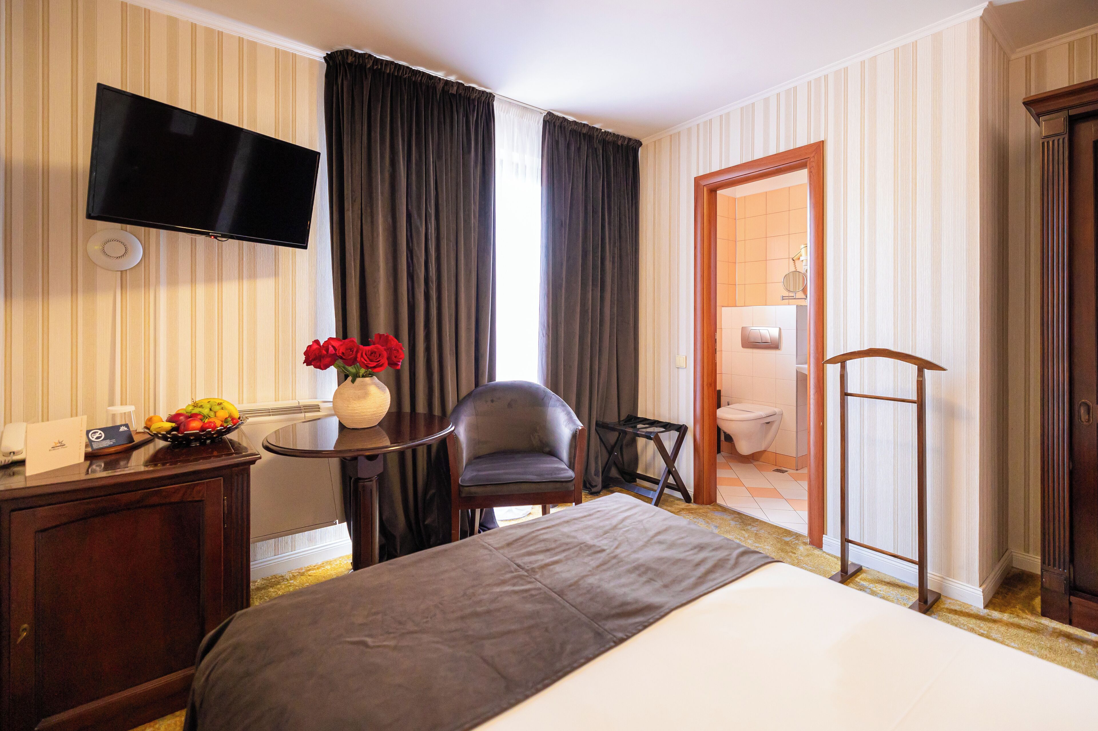 A hotel room with a bed, a desk with a fruit bowl, a chair, a TV, and a bathroom visible through an open door.