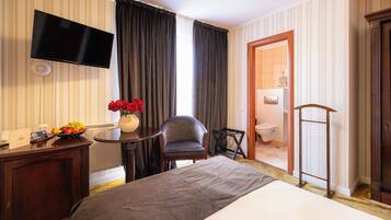 A hotel room with a bed, a desk with a fruit bowl, a chair, a TV, and a bathroom visible through an open door.