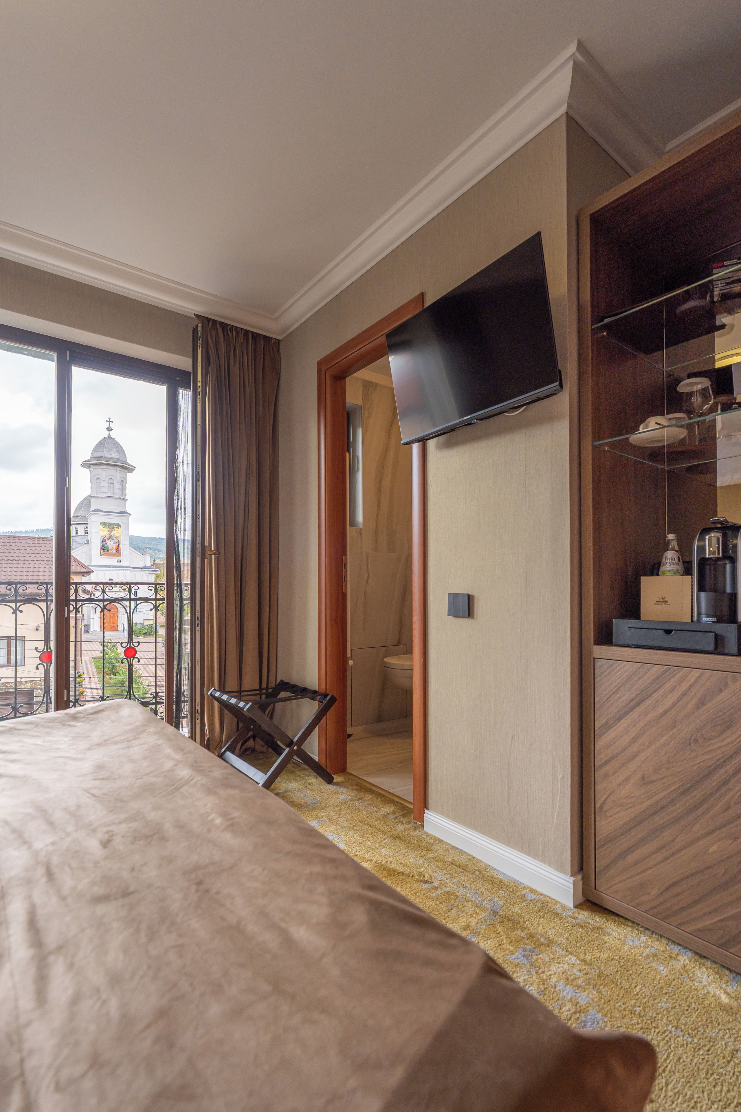 A hotel room with a large bed, a television mounted on the wall, a small table with a bottle, and a view of a church tower through the window.