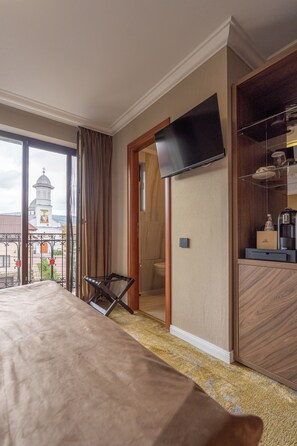 Una habitación de hotel con una cama grande, un televisor montado en la pared, una mesita con una botella y vistas a una torre de iglesia por la ventana.
