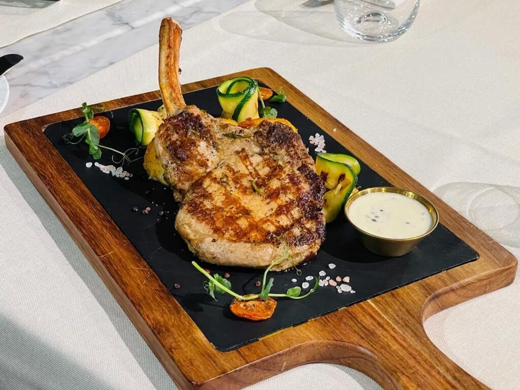 A plated dish with a grilled meat, vegetables, and a small bowl of sauce on a wooden board.