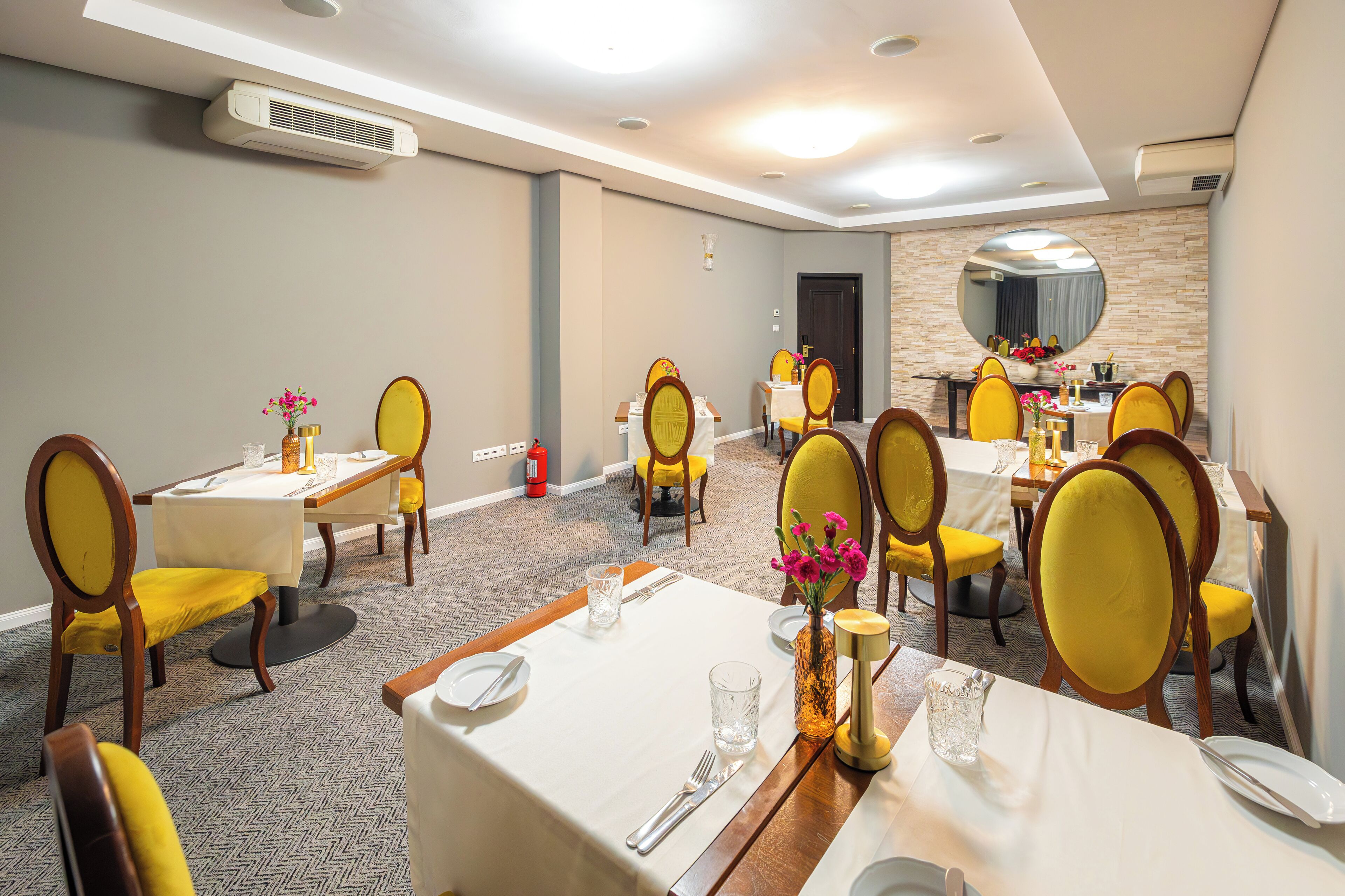 A hotel dining area with multiple tables and yellow chairs, air conditioning units on the walls, and a mirror on the back wall.