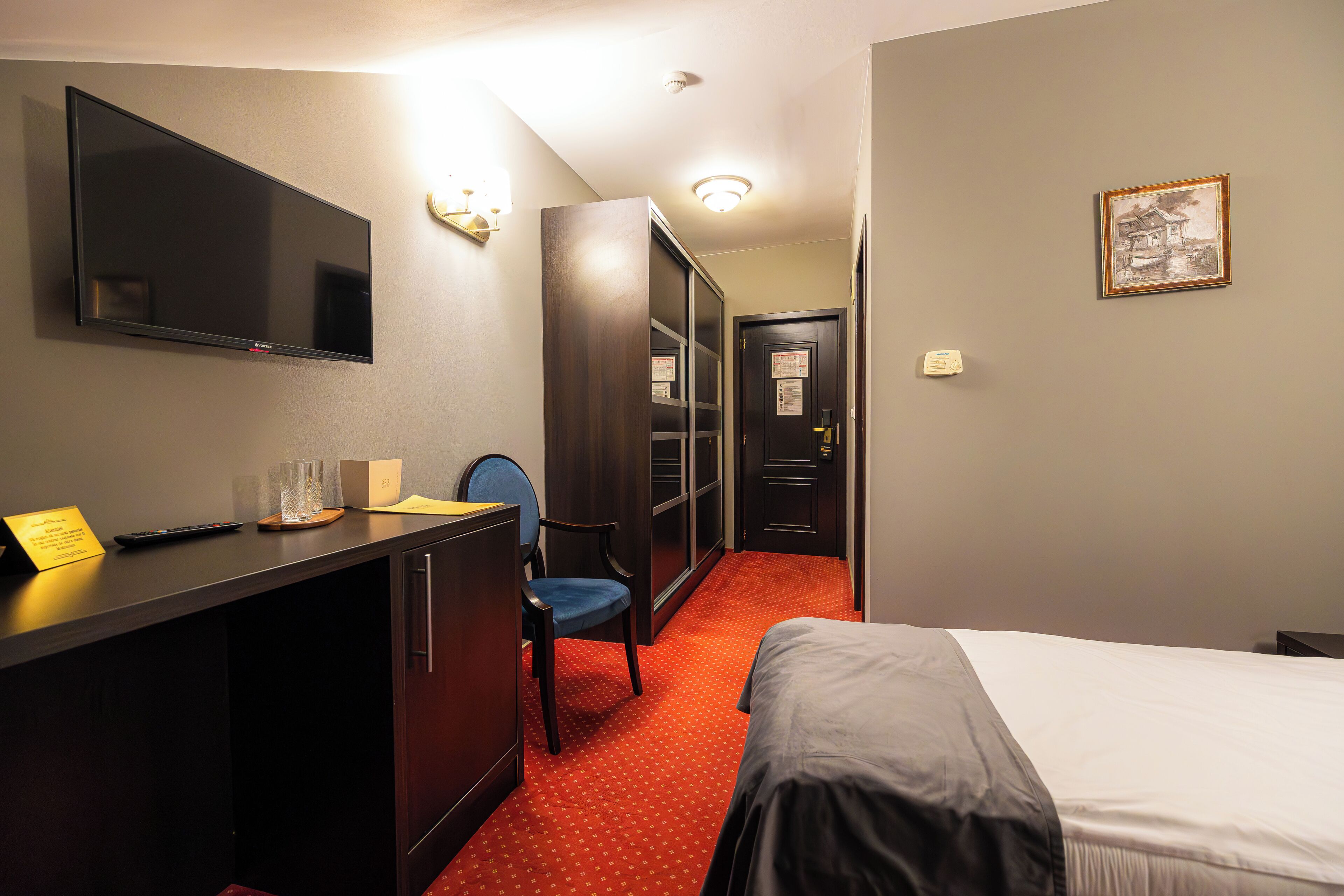 Quarto de hotel com uma cama, uma escrivaninha com computador, uma cadeira, uma televisão e uma porta que dá para o banheiro.