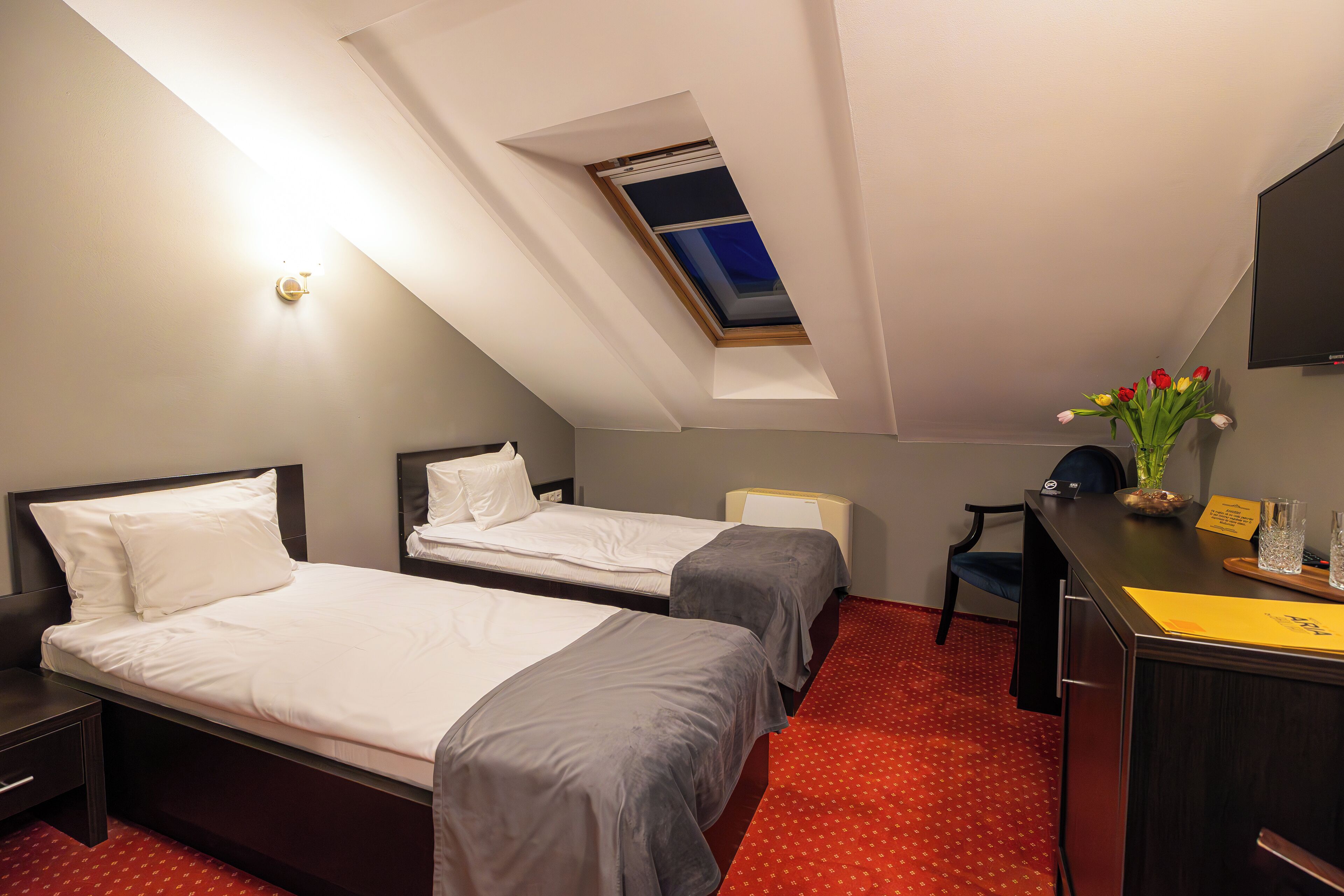 Habitación de hotel con dos camas, un escritorio, una silla, una mesita con un florero y un tragaluz.
