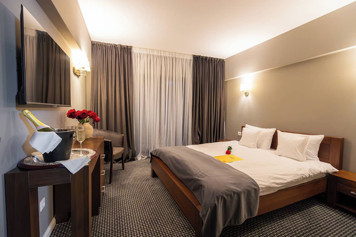 Una camera d'albergo con un letto, una scrivania con una lampada, una bottiglia di champagne e un vaso con fiori rossi.