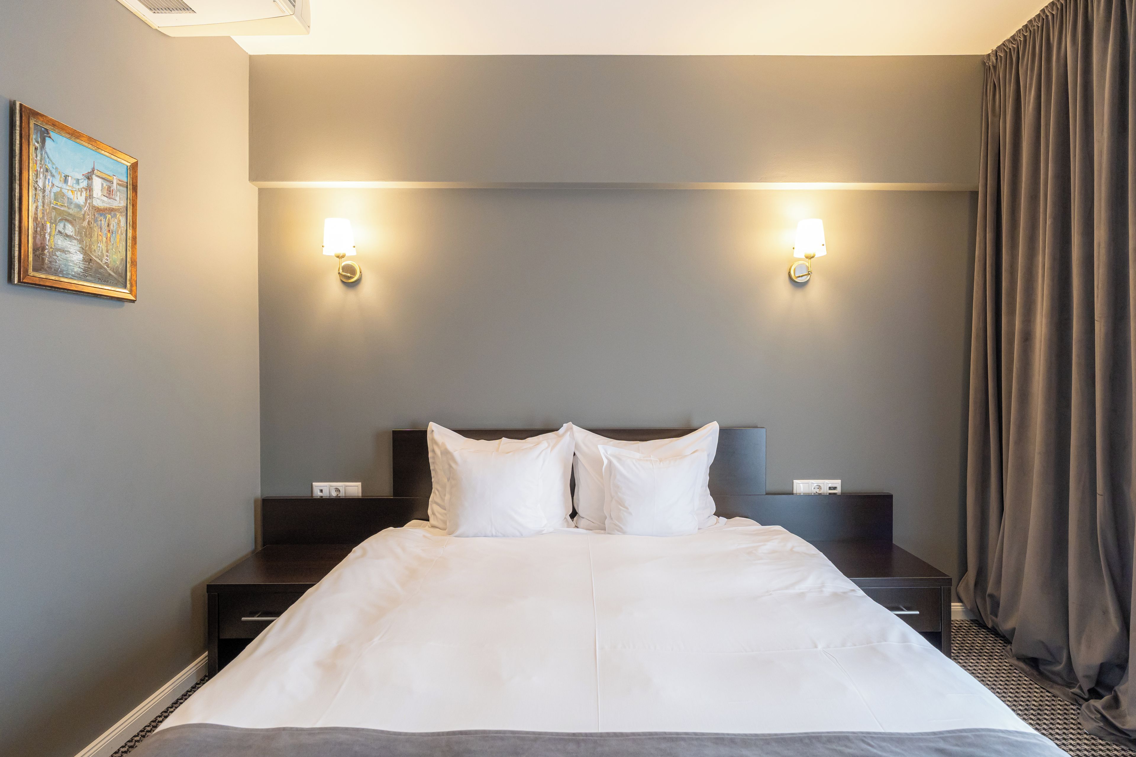 Una camera d'albergo con un letto, due comodini, una testiera, un quadro appeso e una finestra con tende.