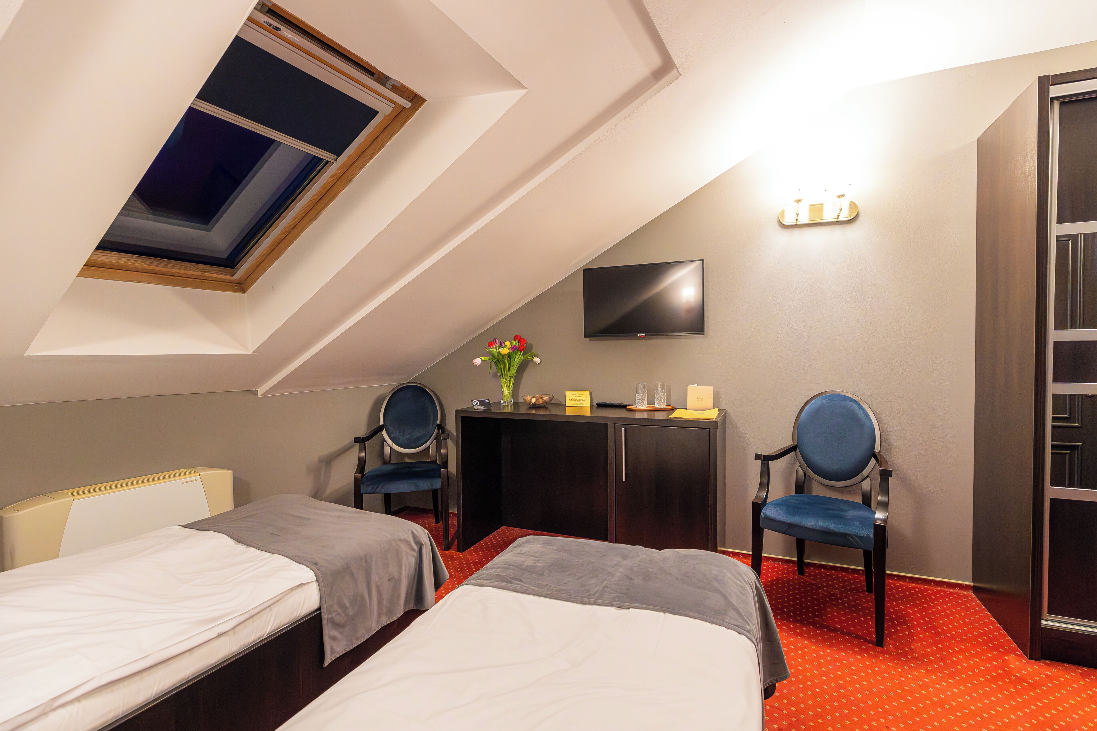 A hotel room with two beds, a desk, a chair, a TV, and a skylight.