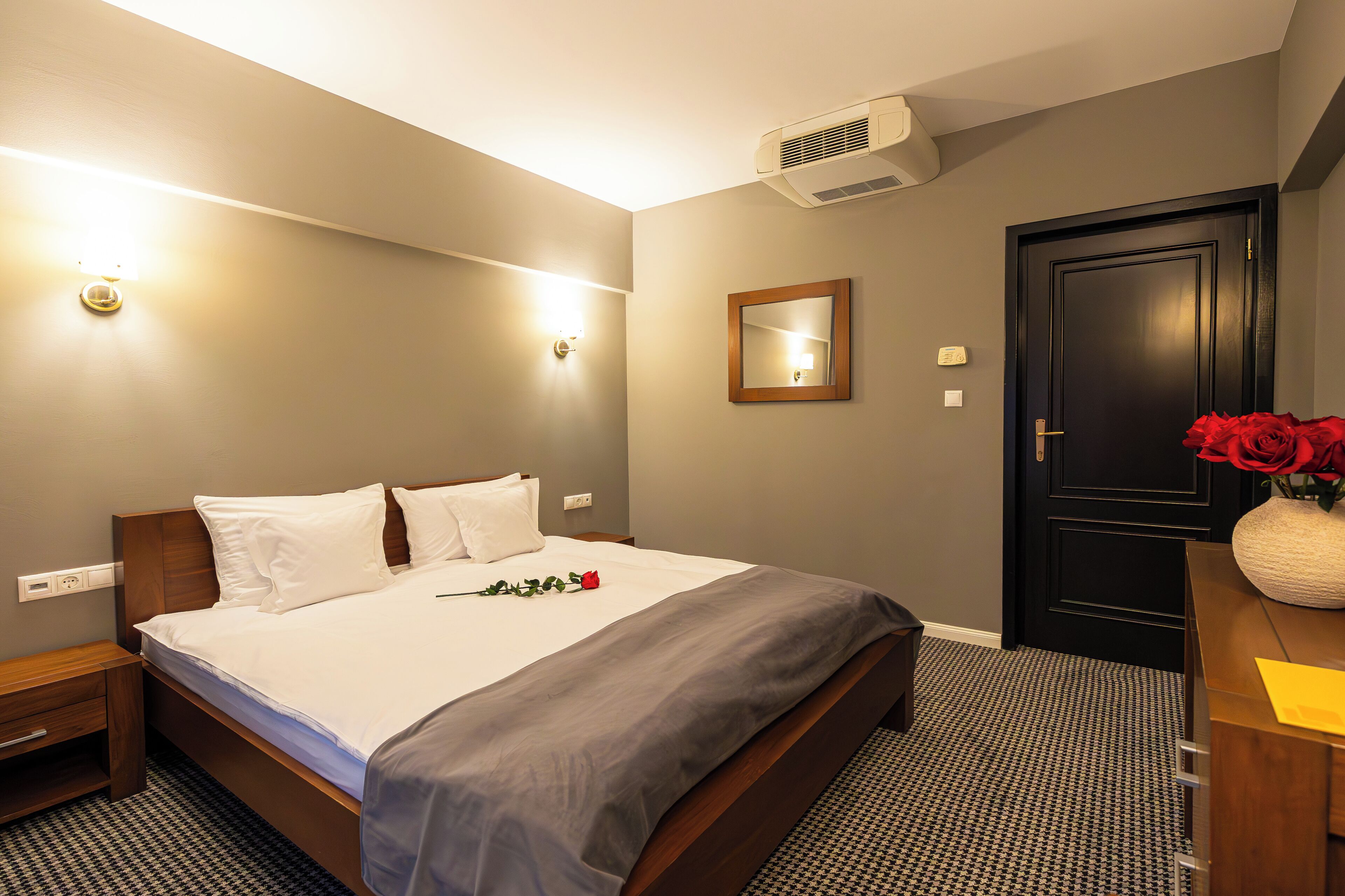A hotel room with a bed, bedside table, and a dresser with a vase of red roses.