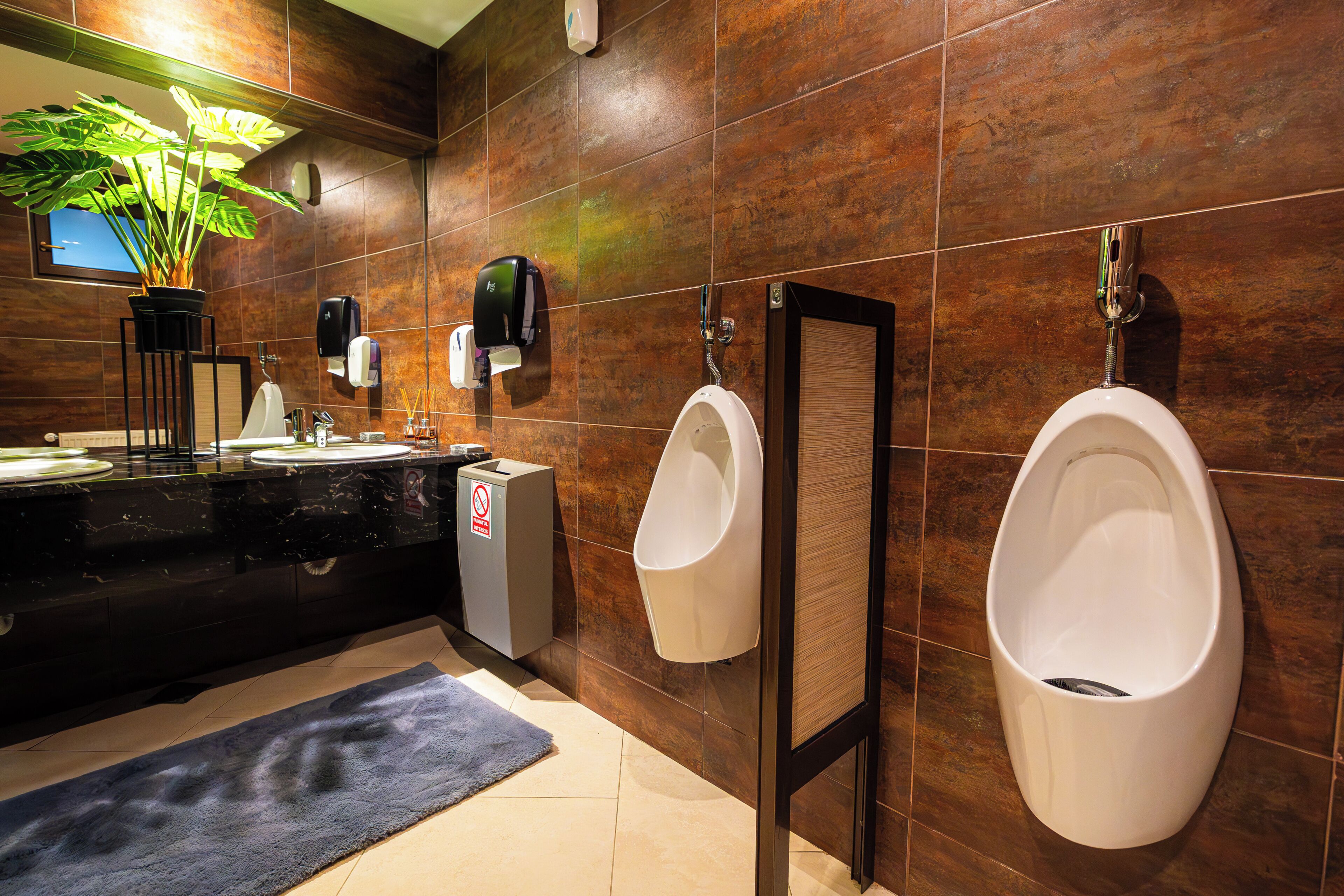 A modern bathroom with a black countertop, two urinals, a hand dryer, a trash can, and a potted plant.