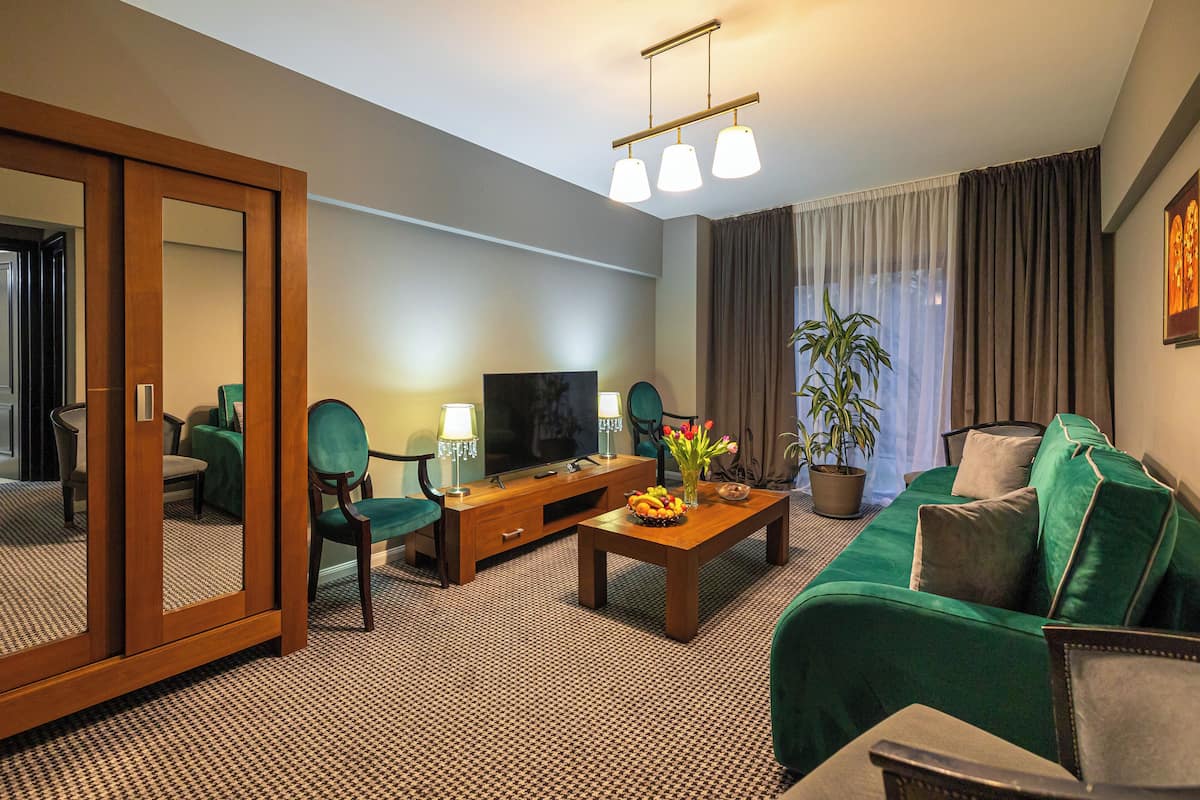 Hotellihuone, jossa on suuri peili, vihreä sohva, puinen sohvapöytä, jossa on hedelmiä, televisio ja ruukussa oleva kasvi.