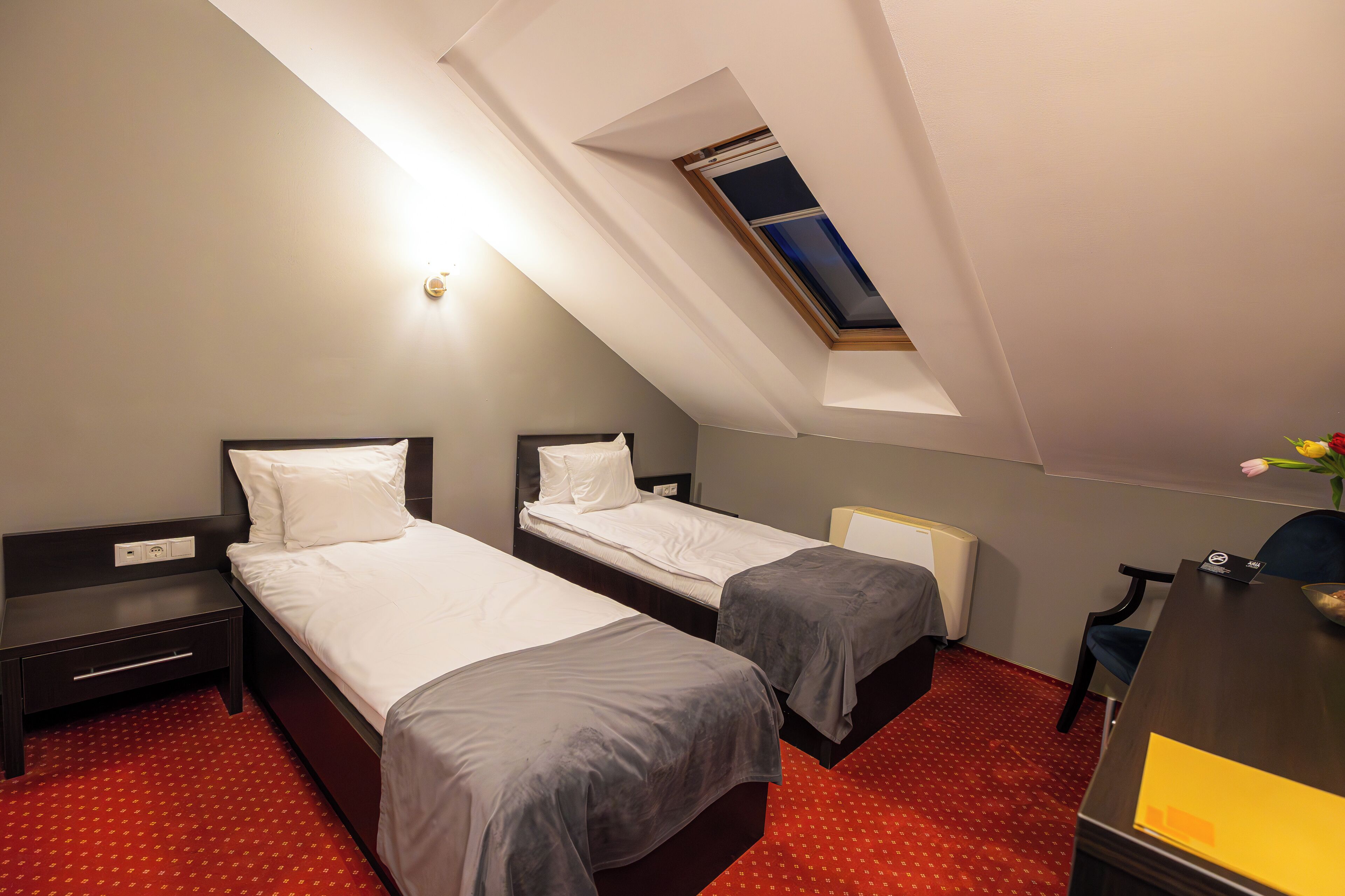 A hotel room with two single beds, a desk, a chair, and a skylight.