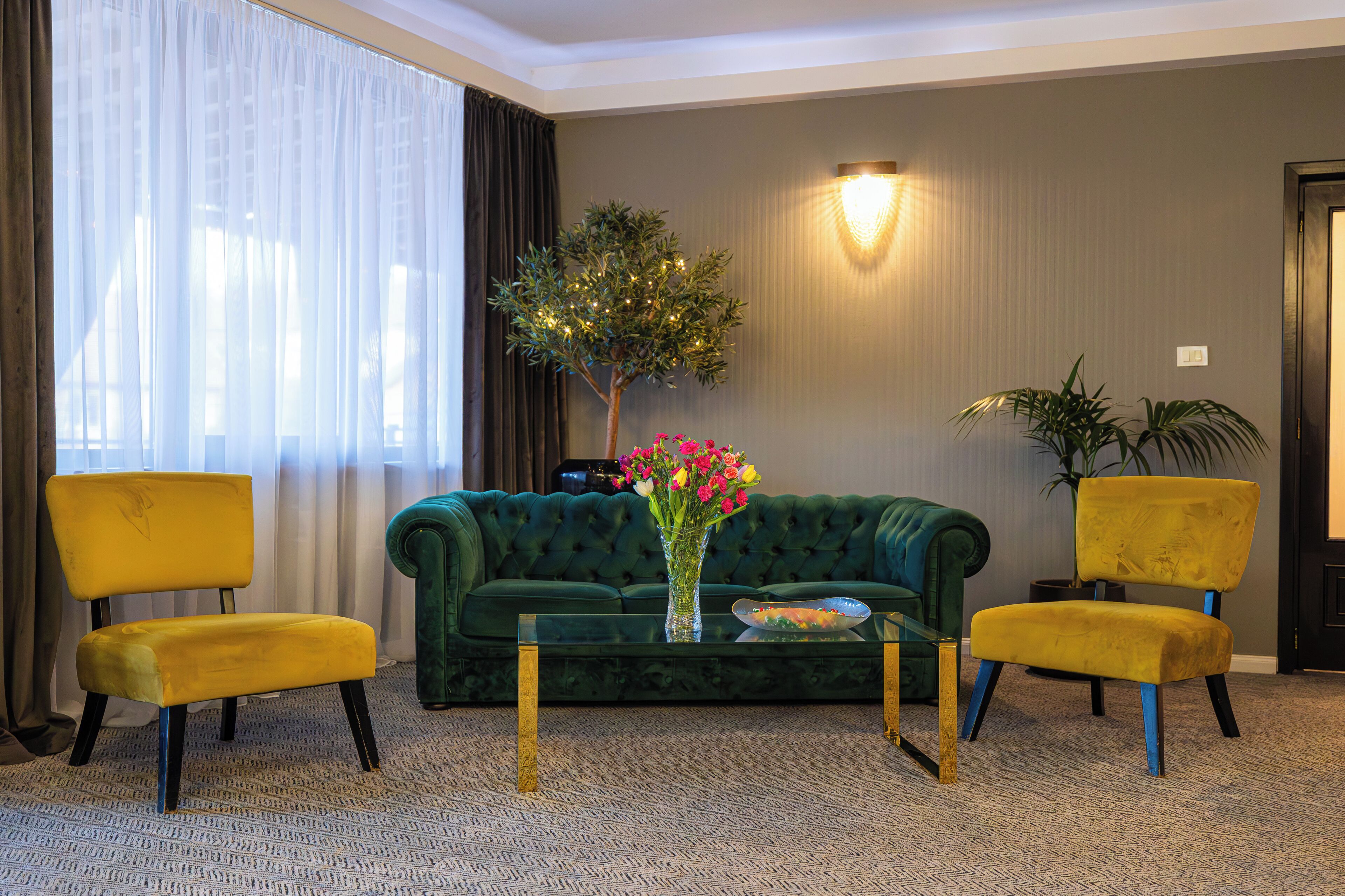 Quarto com sofá verde, duas cadeiras amarelas, mesa de centro de vidro e um vaso com flores.