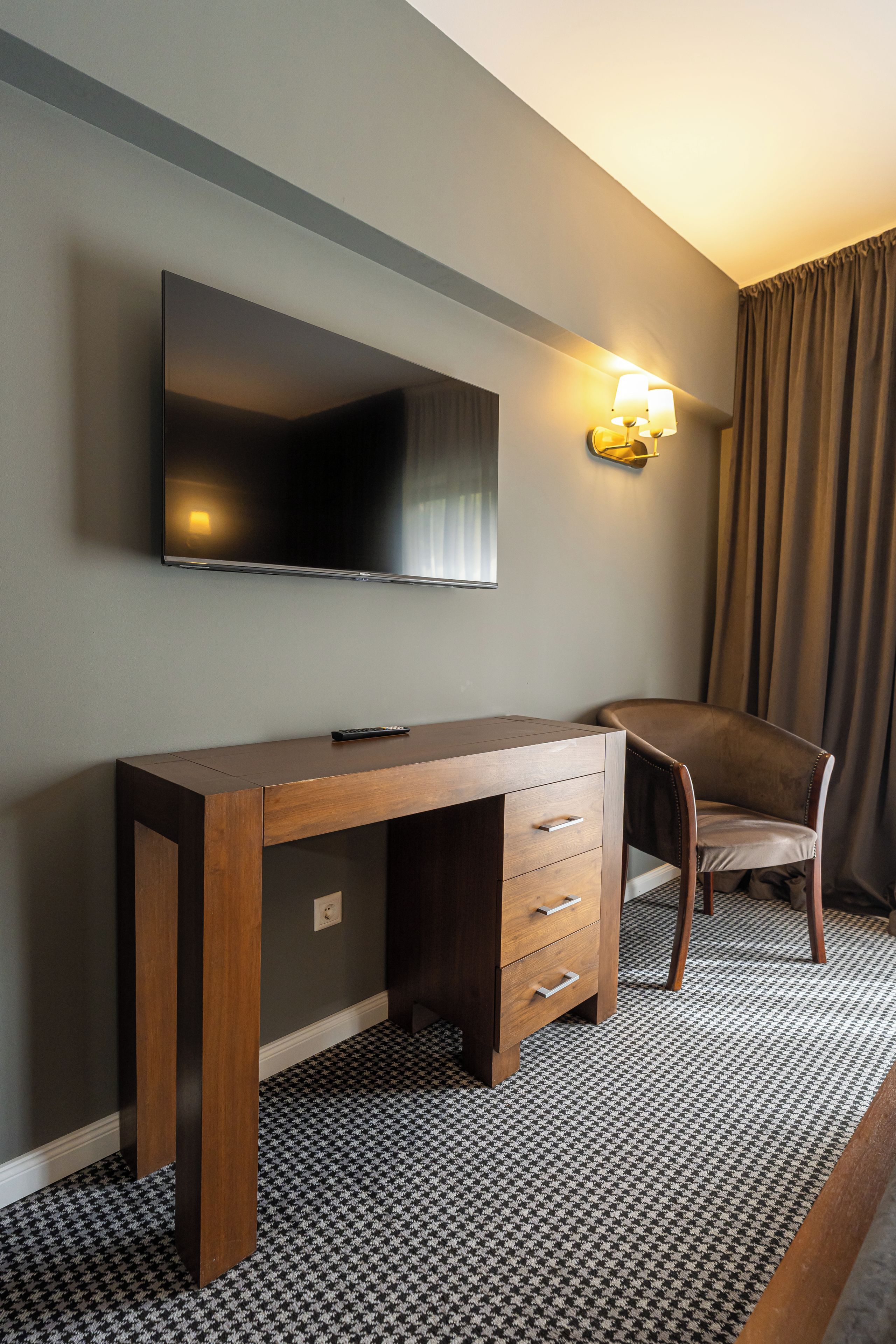 A hotel room with a wooden desk, a flat-screen TV mounted on the wall, a chair, and a patterned carpet.