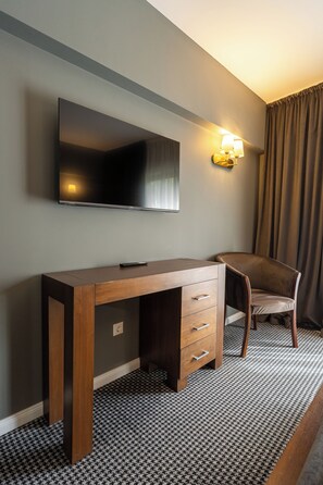 Hotellihuone, jossa on puinen työpöytä, seinälle asennettu litteänäyttöinen televisio, tuoli ja kuviollinen matto.