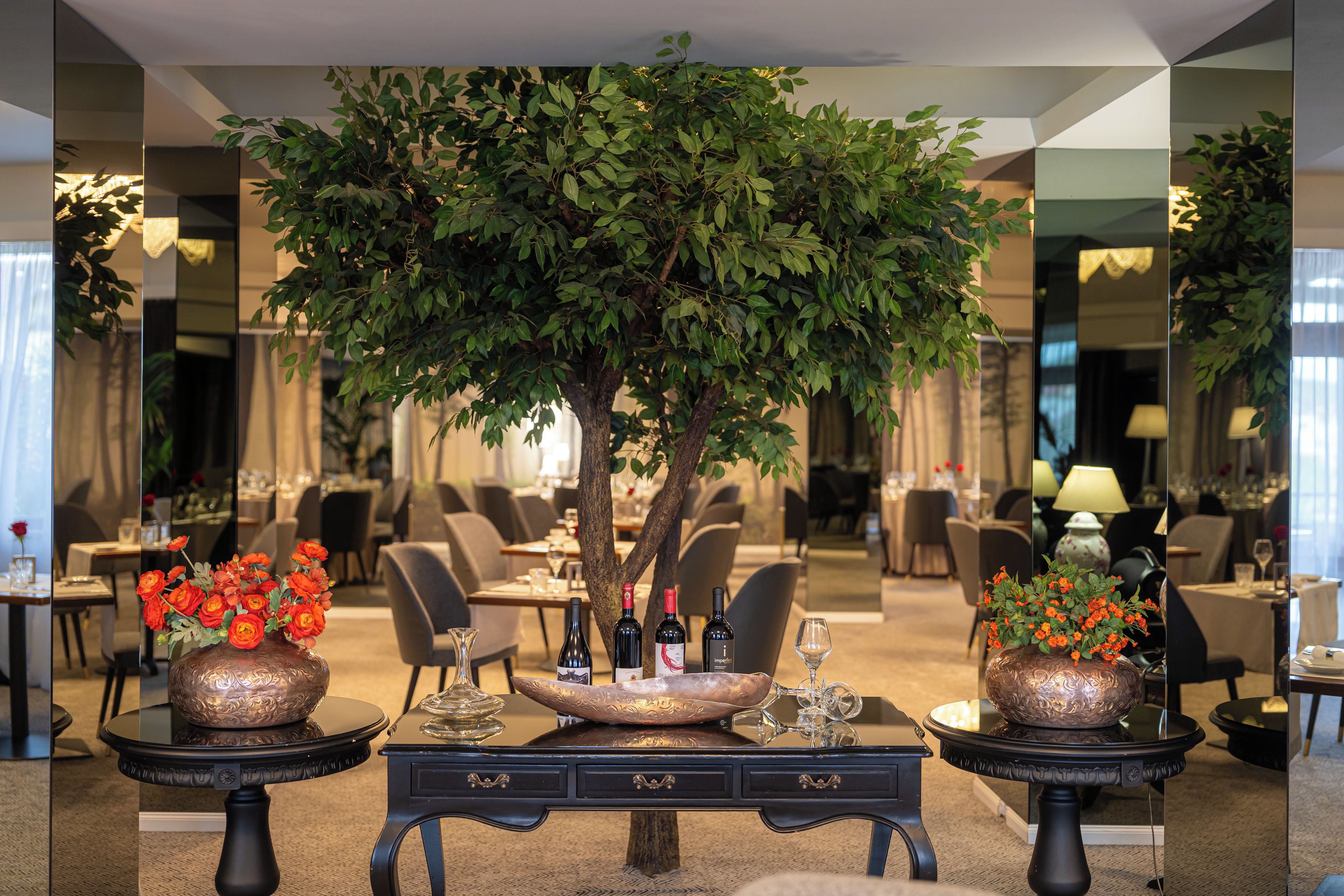 A hotel lobby with a central artificial tree, two side tables with vases of flowers, and a mirrored wall reflecting the dining area.