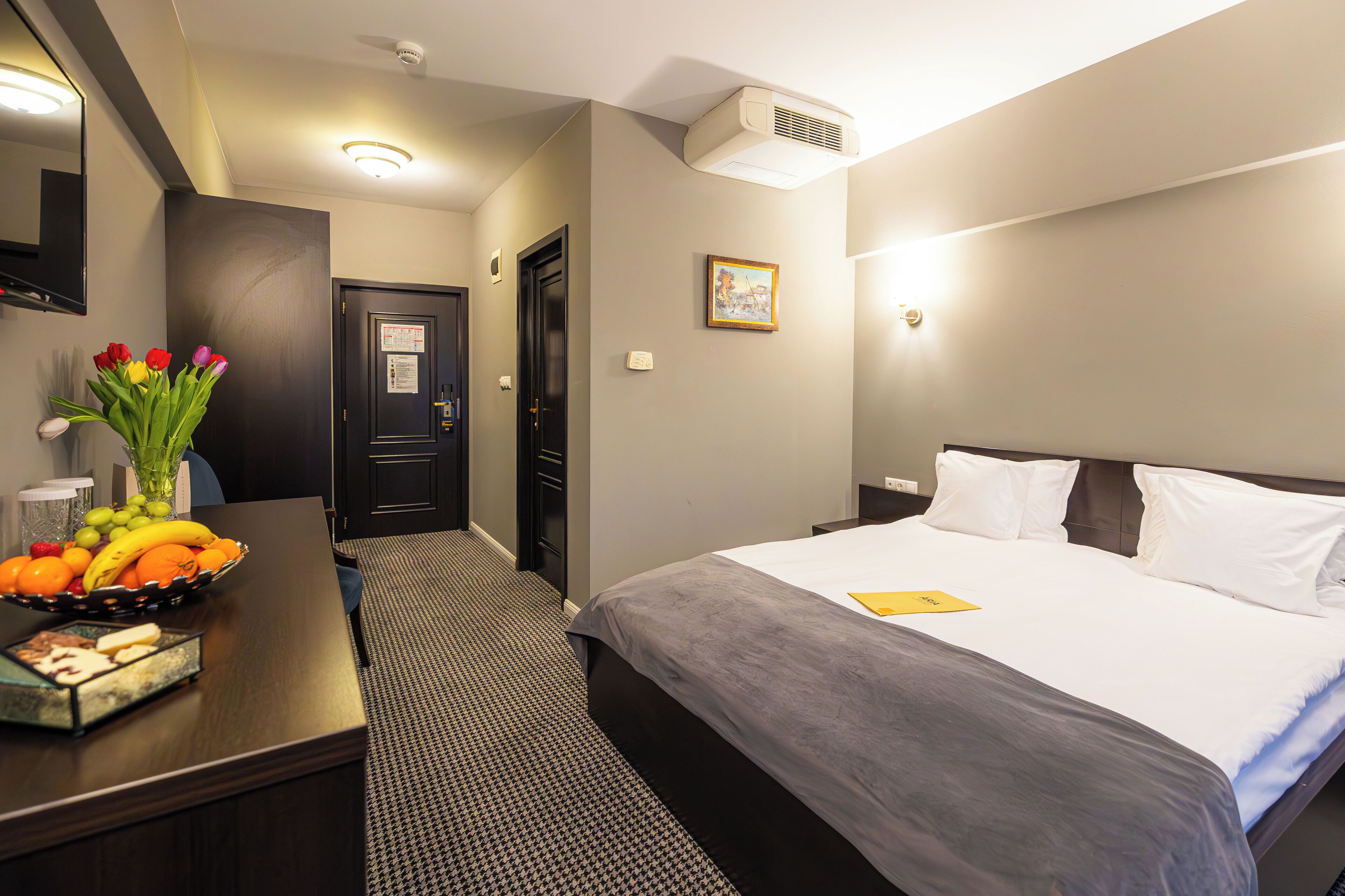Een hotelkamer met een bed, een bureau met een schaal fruit, een vaas met bloemen en een ingelijst foto aan de muur.