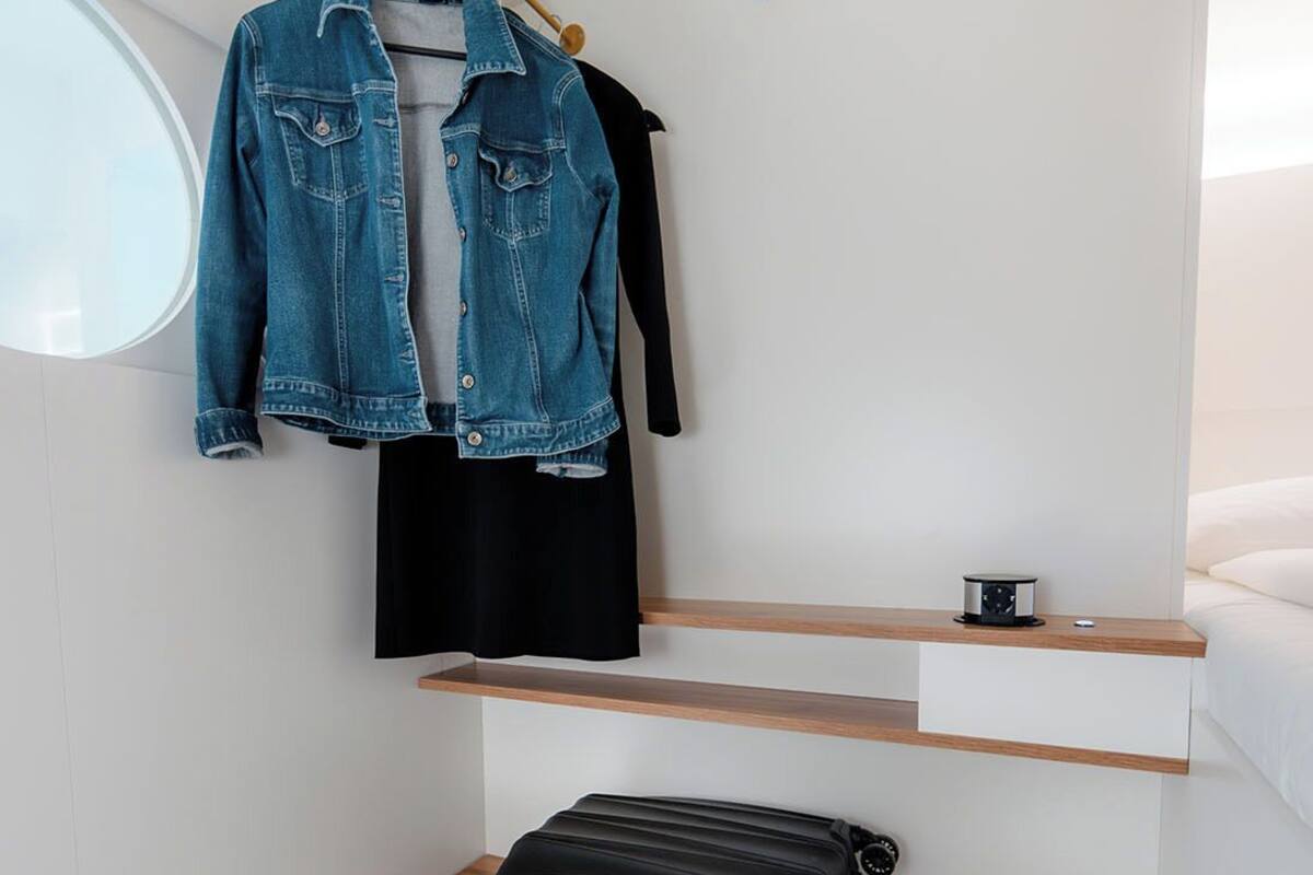 A compact, modern bathroom with a wooden vanity, a black suitcase, a denim jacket, and a black shirt hanging on a wooden rack.