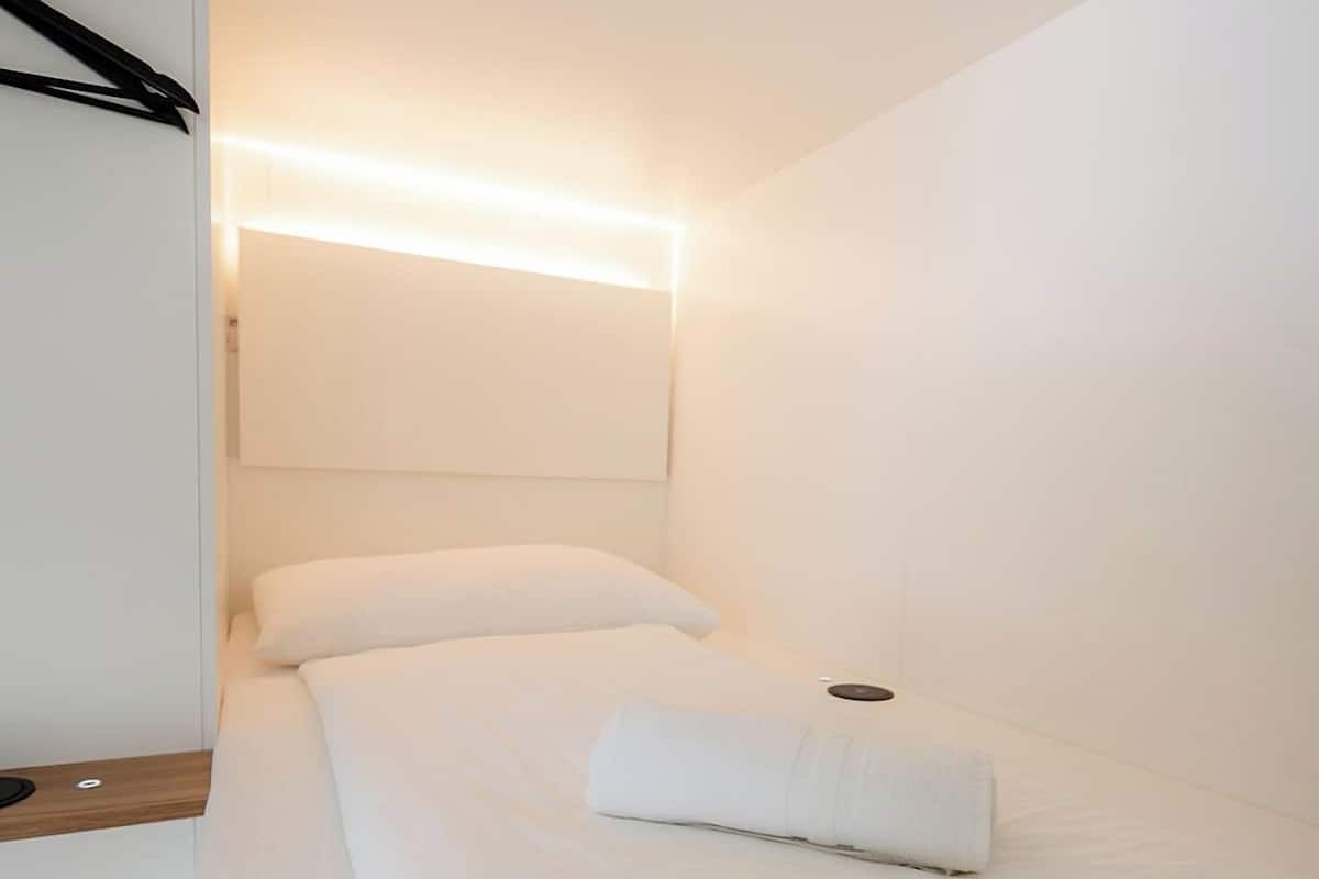 A single bed with white bedding and a neatly folded towel, a bedside table, and a wall-mounted light fixture.