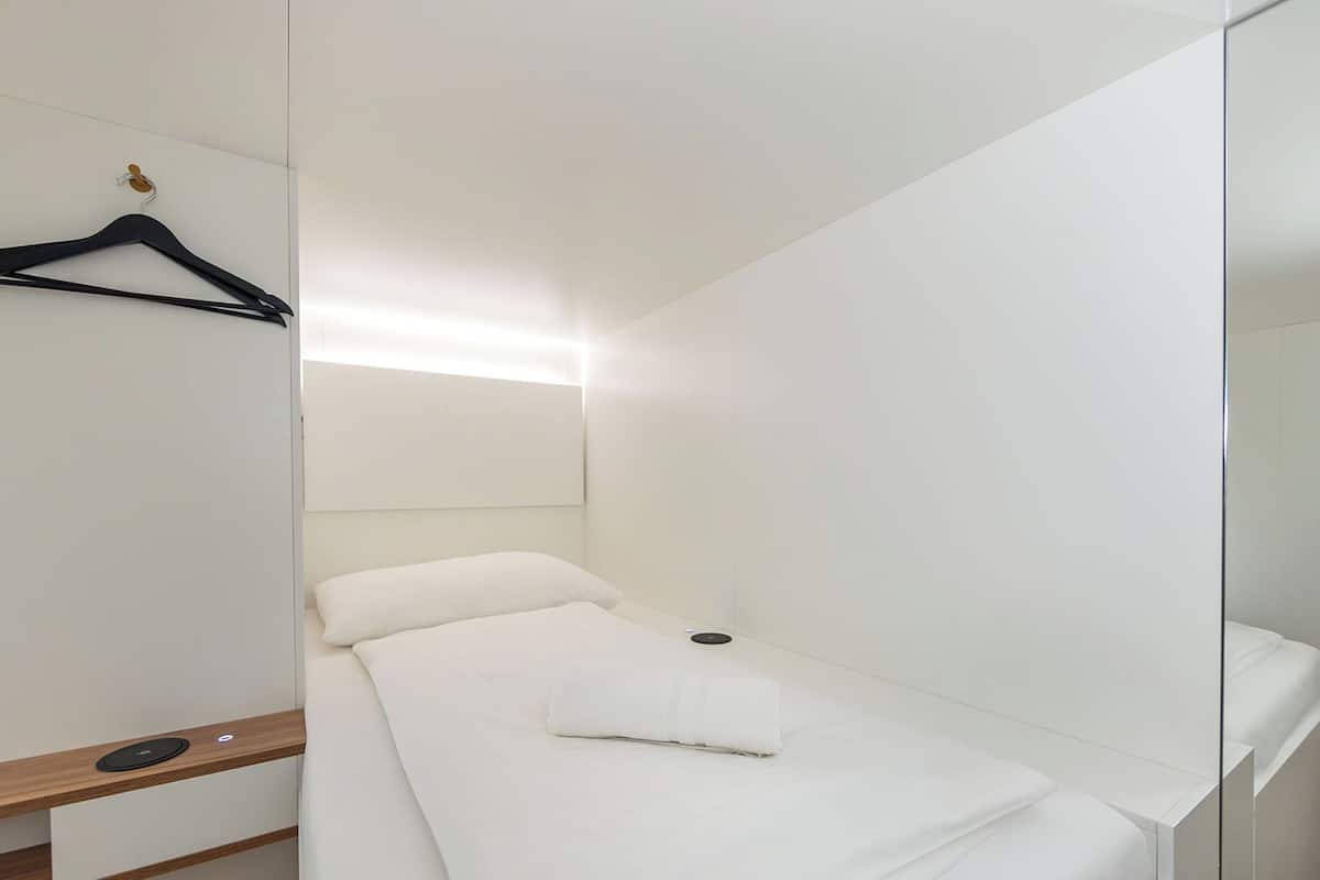 A small, single-bed room with a wooden bedside table, a hanging black hanger, and a mirror on the wall.