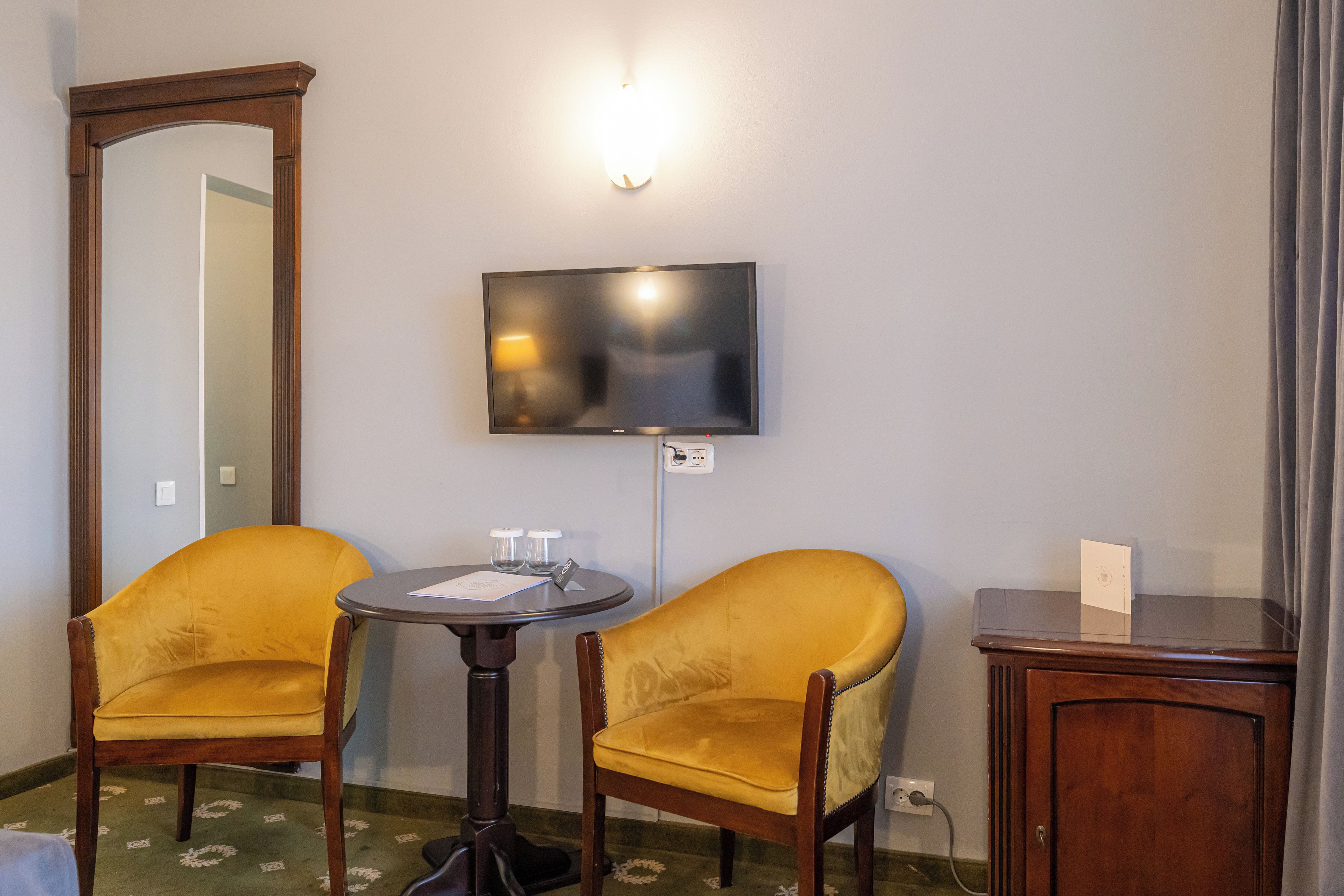 Habitación de hotel con dos sillones amarillos, una mesita redonda, un televisor de pantalla plana montado en la pared y un armario de madera.
