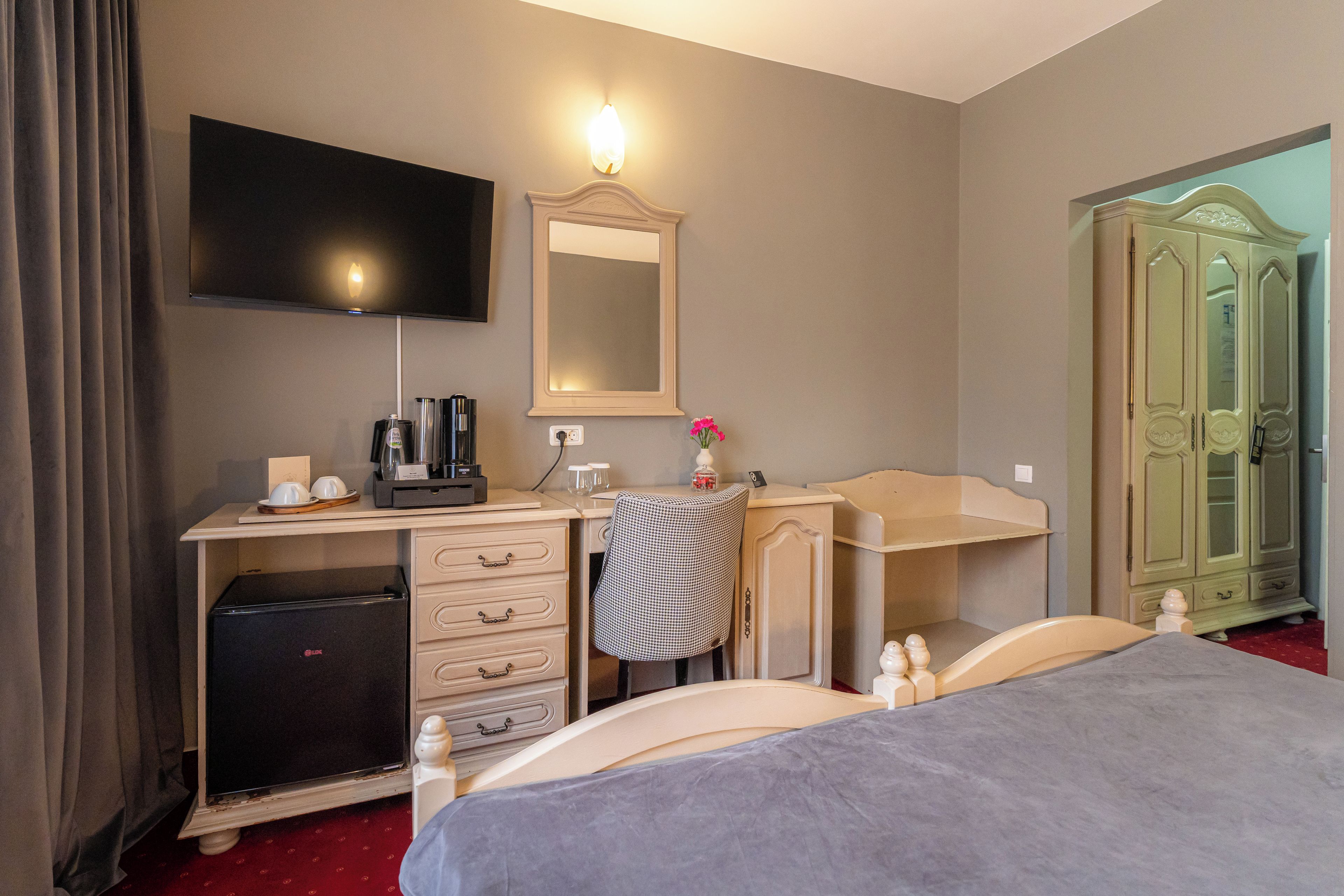 A hotel room with a bed, a desk with a flat-screen TV, a mirror, a dresser, a wardrobe, and a minibar.
