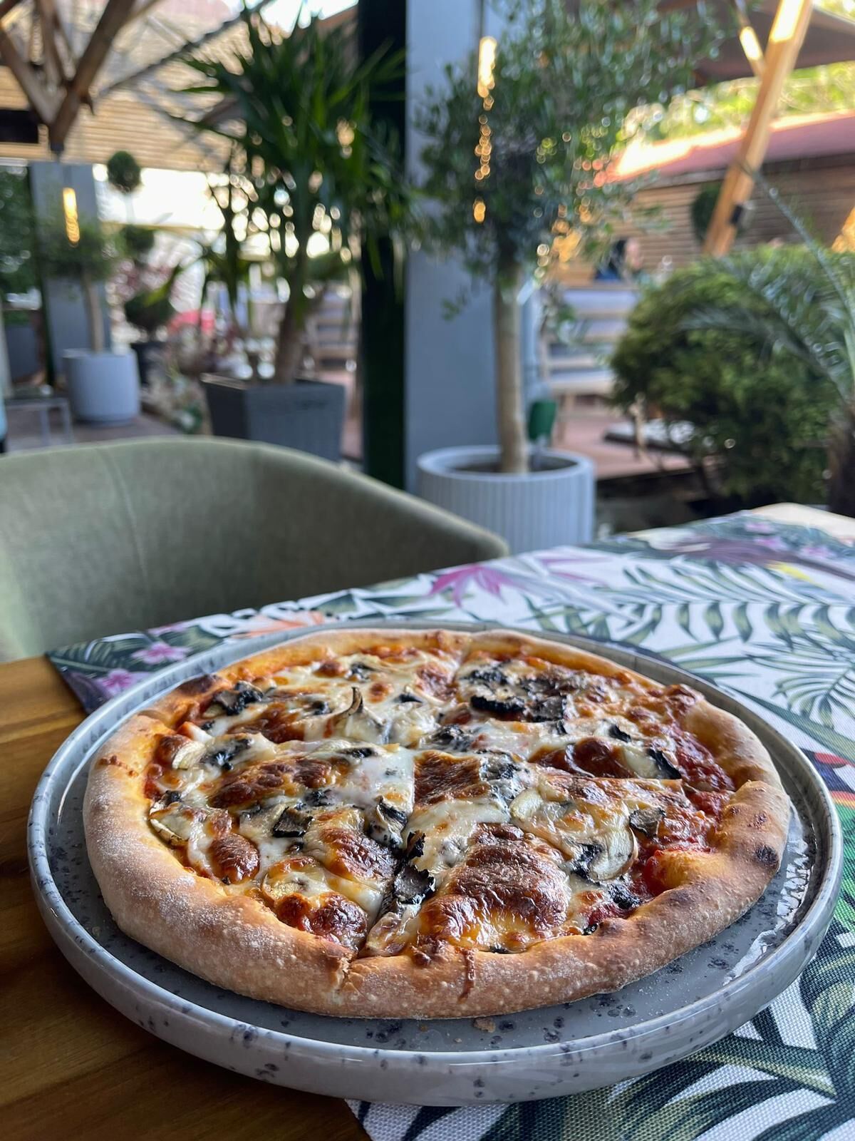 A pizza on a metal tray with a patterned tablecloth, set in an outdoor dining area with chairs and potted plants.