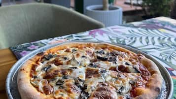 A pizza on a metal tray with a patterned tablecloth, set in an outdoor dining area with chairs and potted plants.
