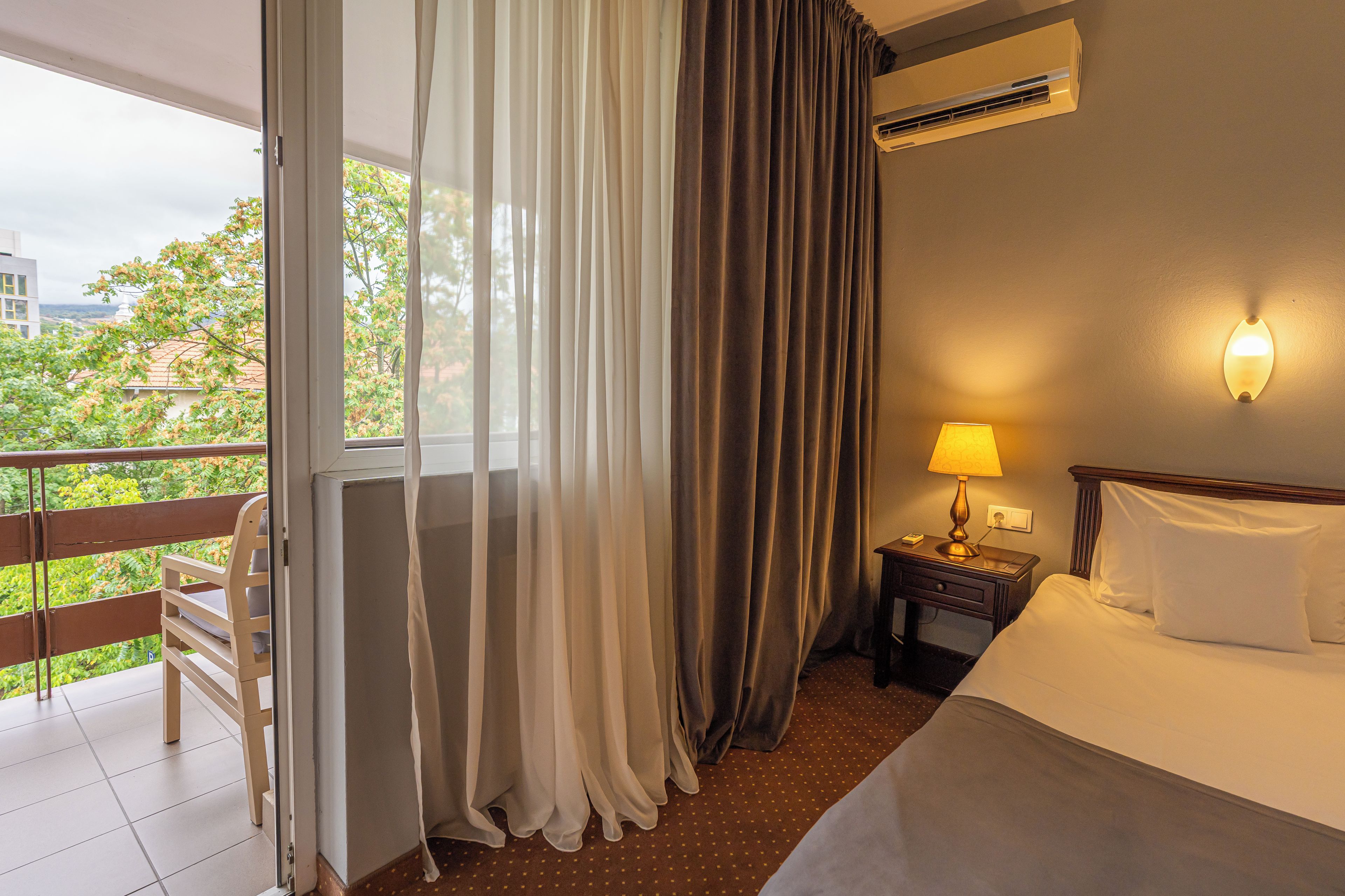 Habitación de hotel con cama, mesita de noche con lámpara, ventana con cortinas y equipo de aire acondicionado.