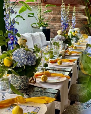 Une longue table à manger, dressée avec des serviettes jaunes, des verres transparents et une variété de fleurs, notamment des hortensias et des fleurs violettes.