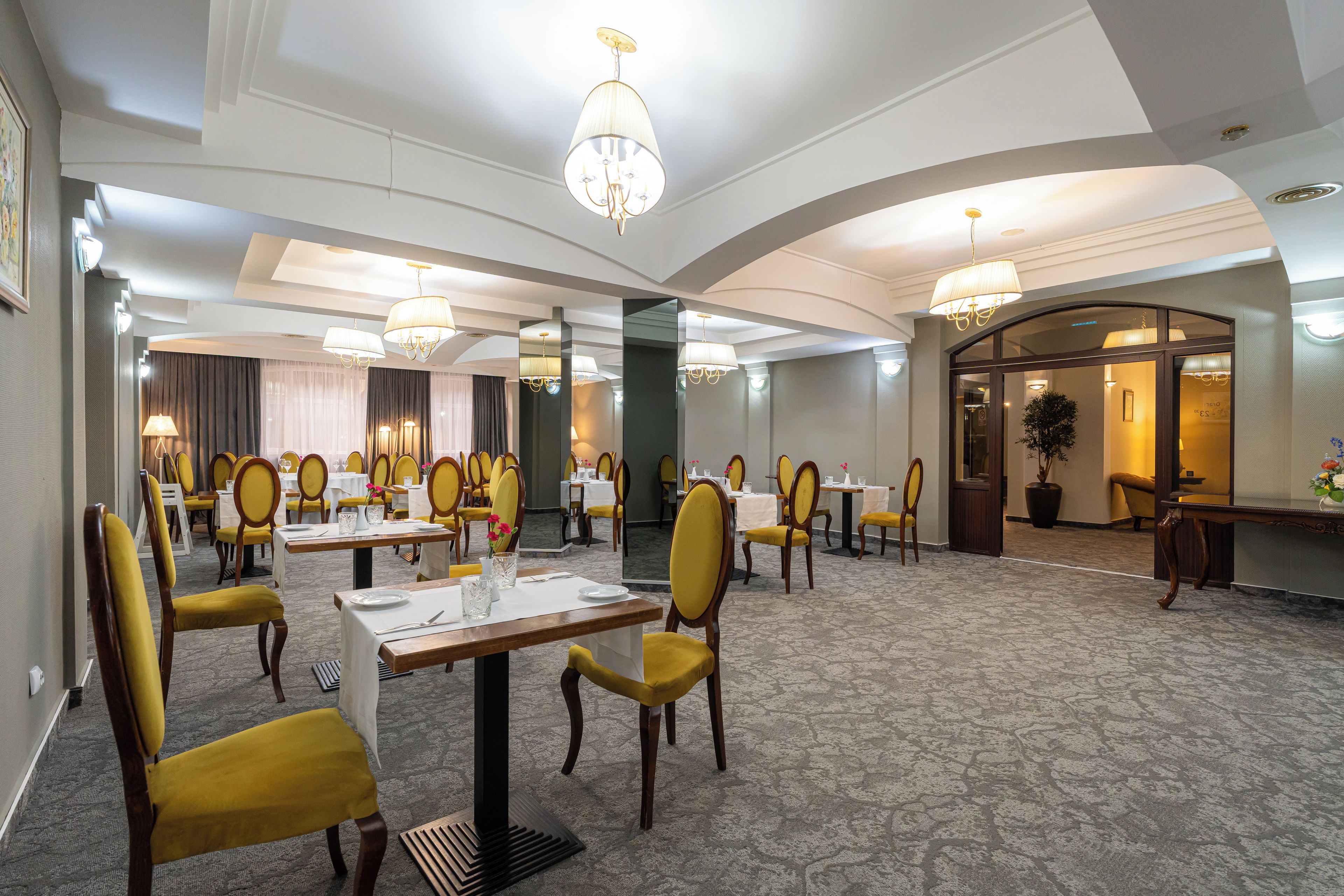 Een restaurantgedeelte in een hotel met meerdere tafels en stoelen, kroonluchters en een vloerkleed.