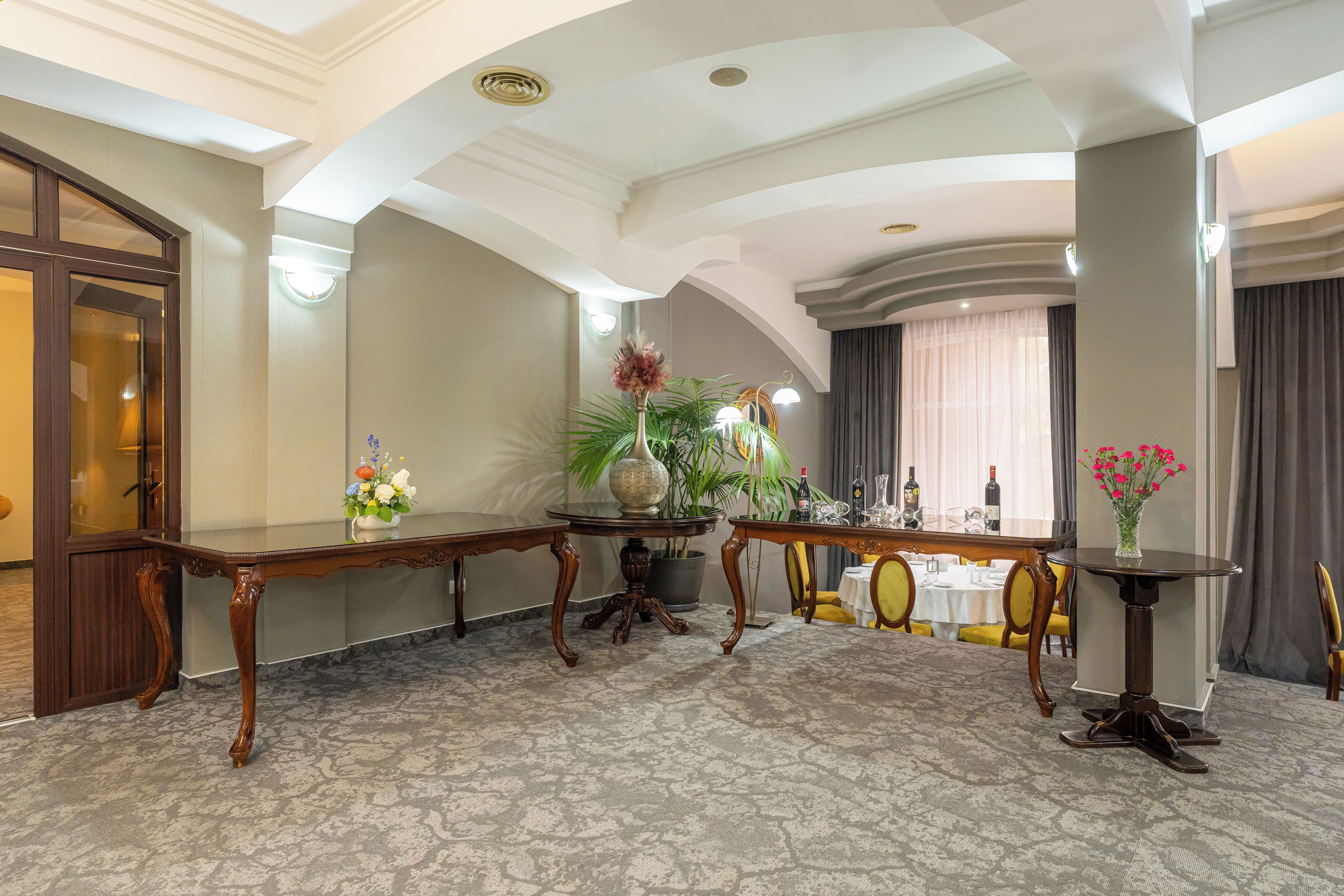A hotel lobby with a marble floor, wooden furniture, a large vase with flowers, and a view of the dining area through a window.
