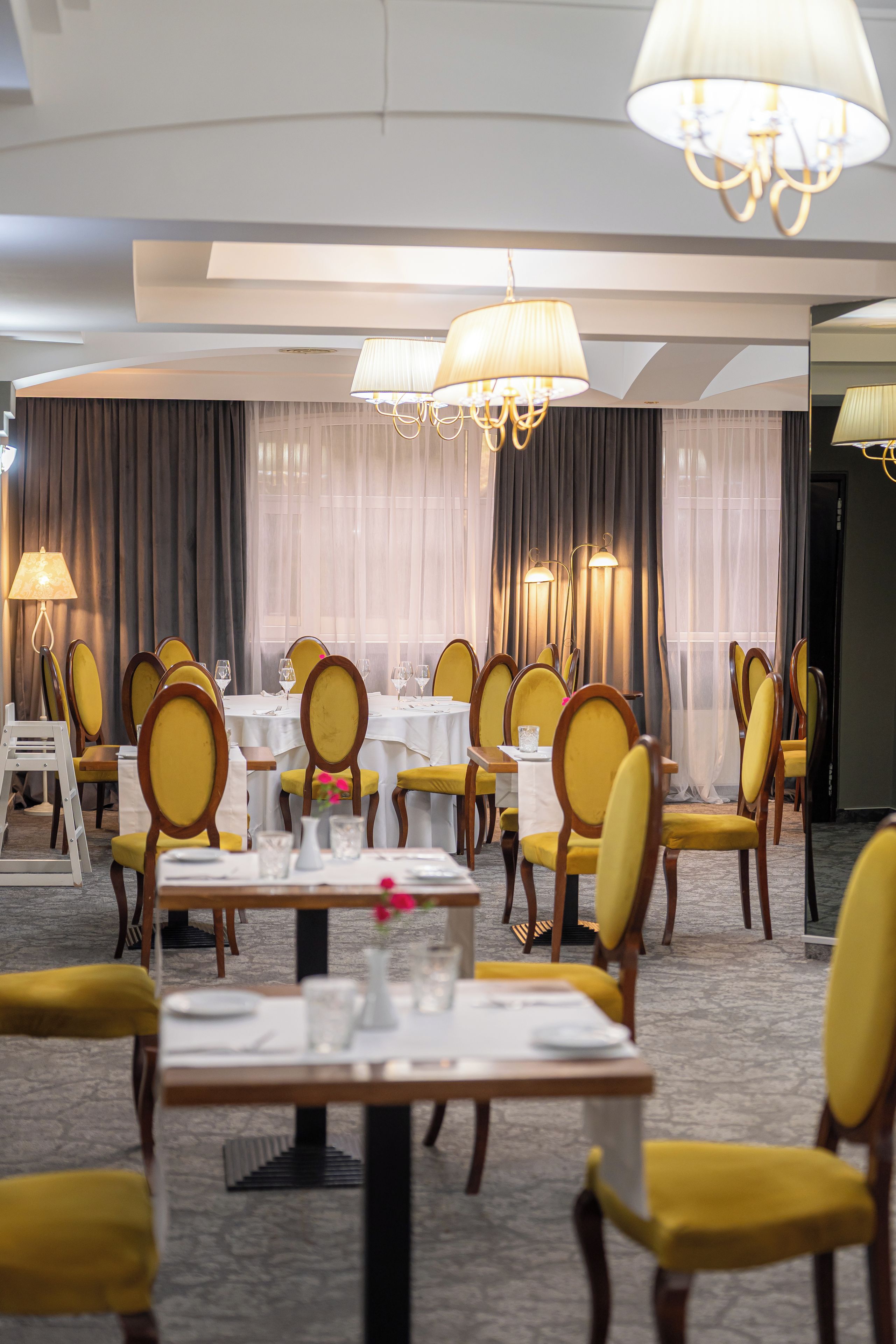 A hotel dining area with tables set for guests, yellow upholstered chairs, and chandeliers.