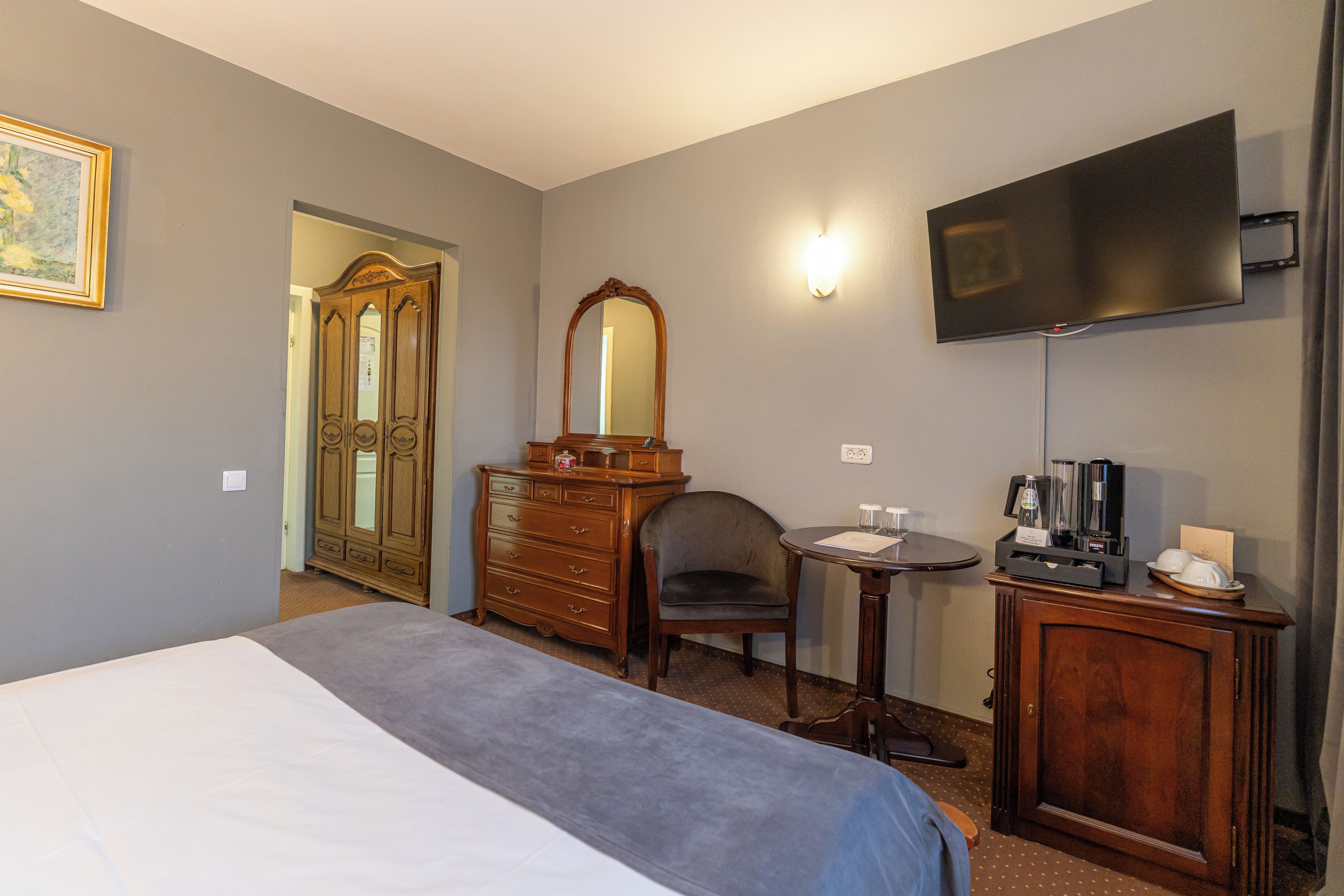 Ein Hotelzimmer mit einem Bett, einem Schminktisch, einem Sessel, einem kleinen Tisch, einem an der Wand befestigten Fernseher und einer Tür zu einem anderen Raum.