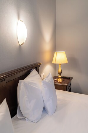 Una cama bien hecha con sábanas y almohadas blancas, una lámpara de noche y un aplique de pared.