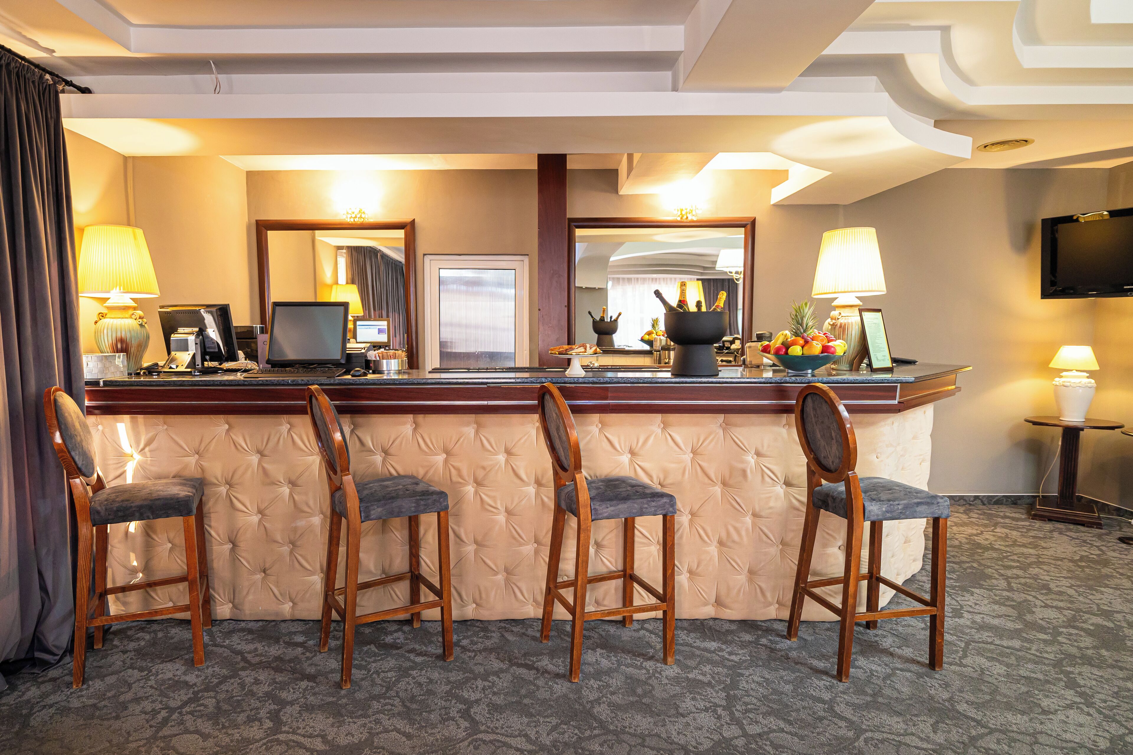 Angolo di un bar dell'hotel con quattro sgabelli in legno, un portatile, una ciotola di frutta e un televisore appeso al muro.