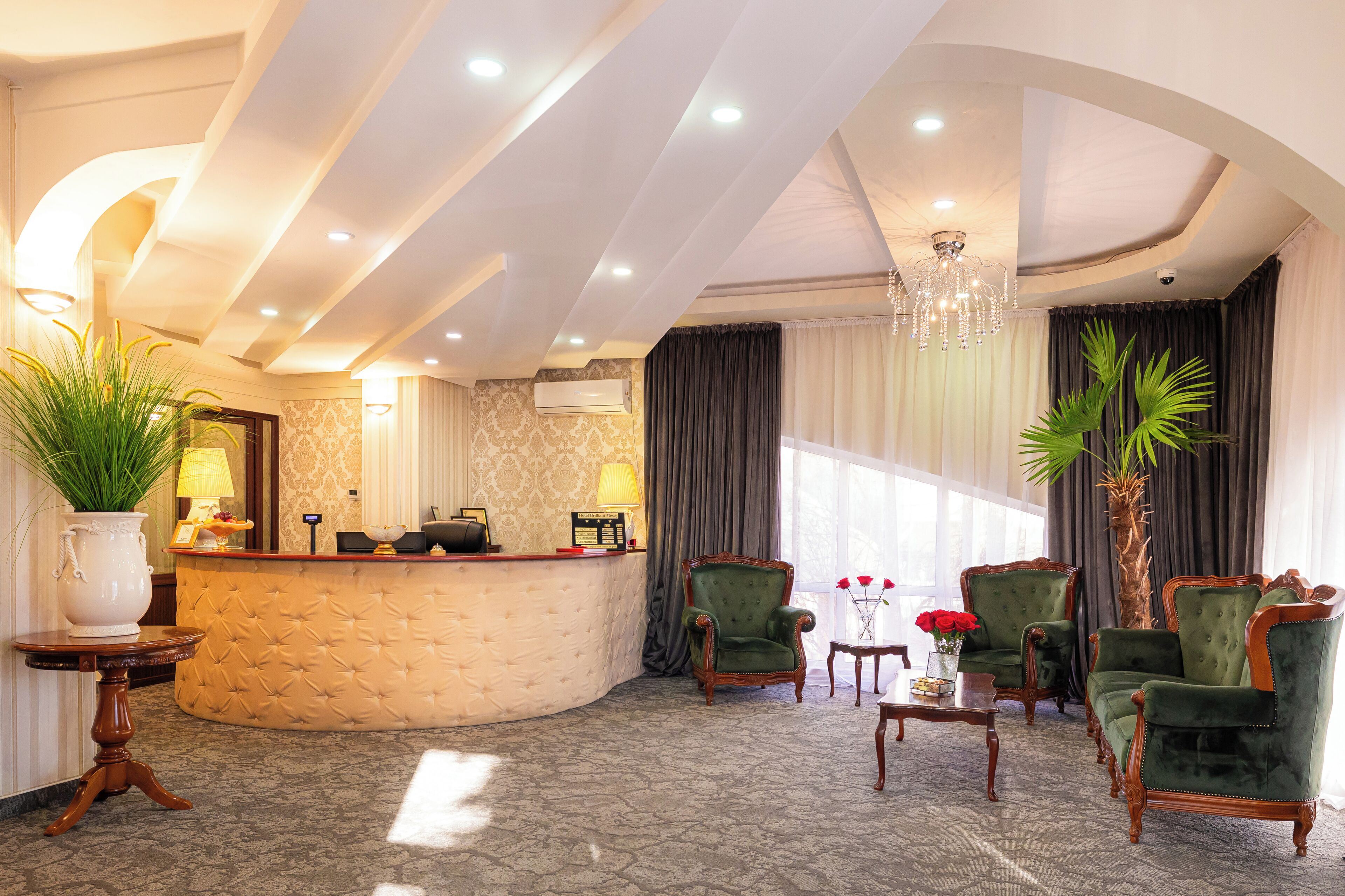 A hotel lobby with a reception desk, green upholstered chairs, a chandelier, and a large potted plant.