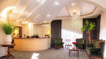 A hotel lobby with a reception desk, green upholstered chairs, a chandelier, and a large potted plant.