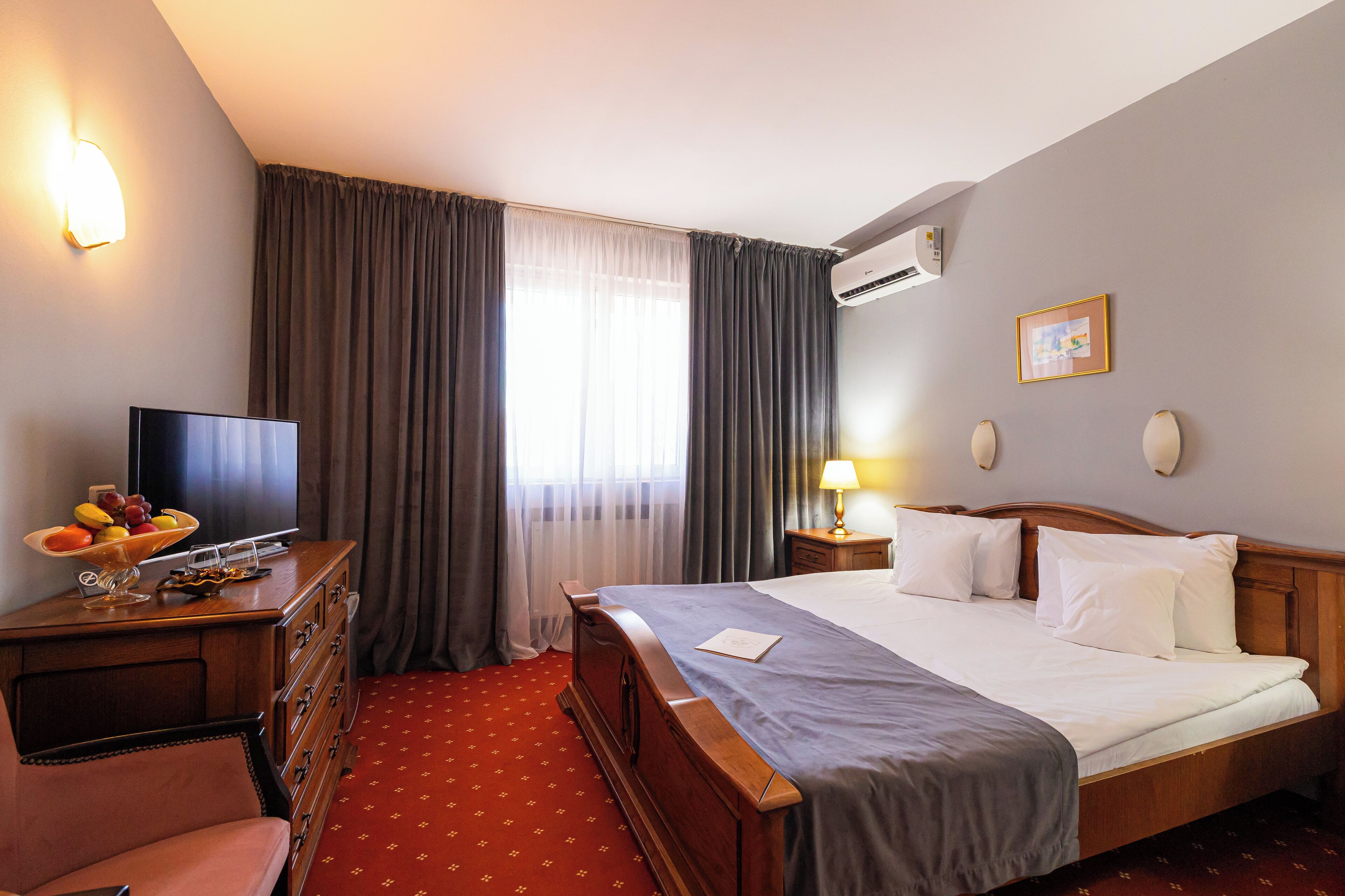 A hotel room with a bed, a TV, a desk with a fruit bowl, and a red carpet.