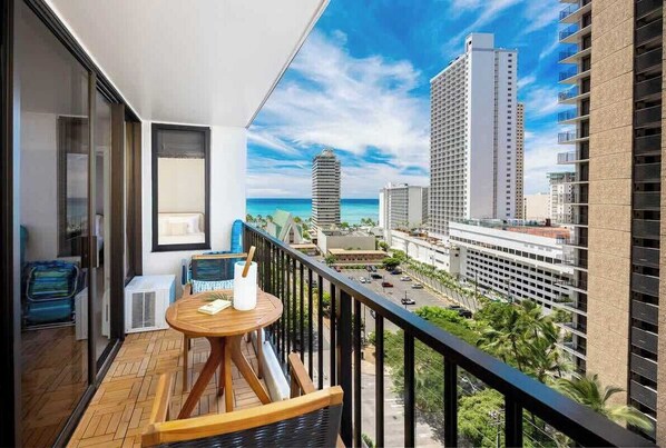 A balcony with a wooden table and chairs, overlooking a cityscape with tall buildings and a clear sky.