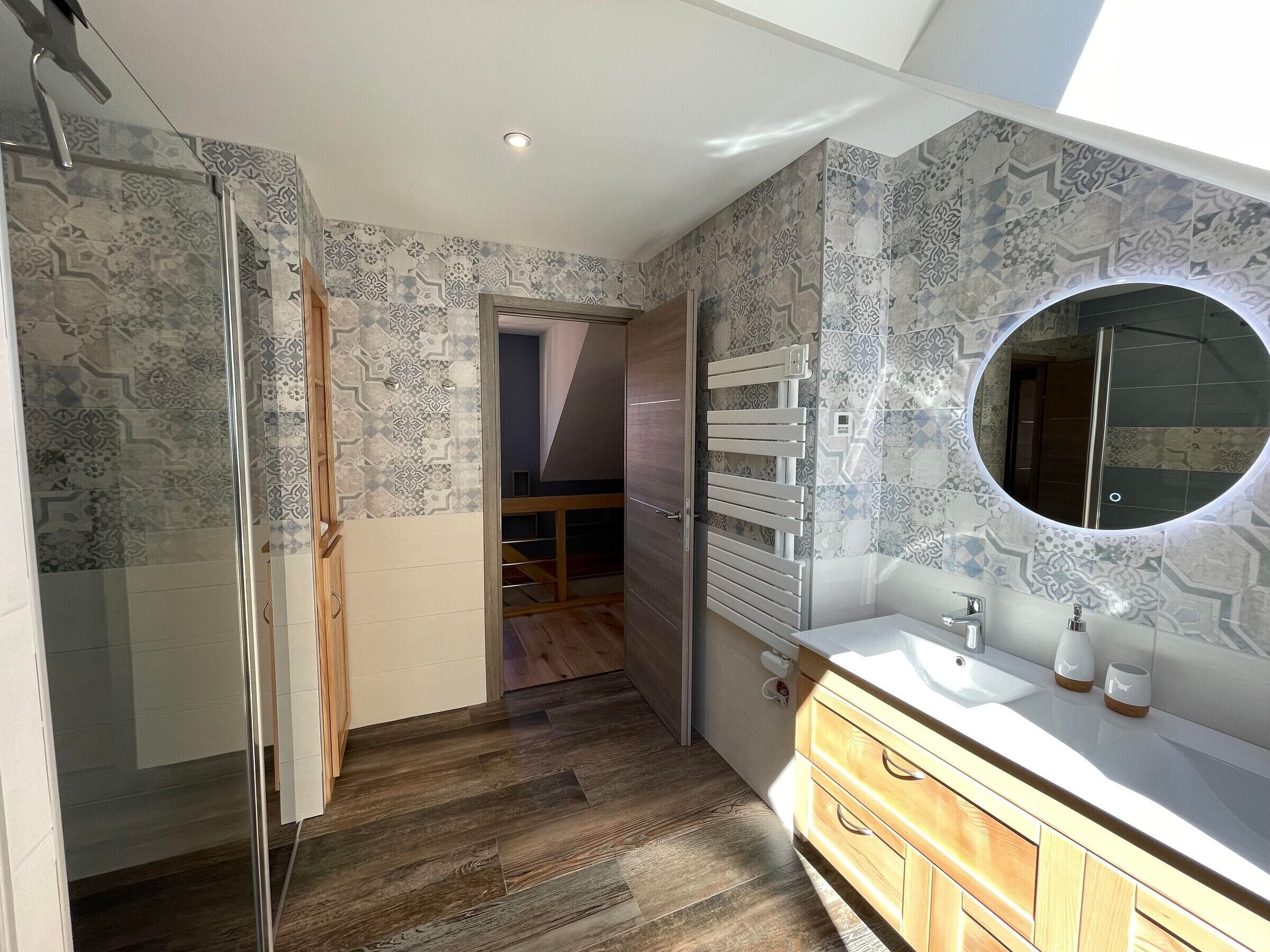 Een moderne badkamer met patroon betegelde vloeren, een glazen douchecabine, houten kastjes en een ronde spiegel.