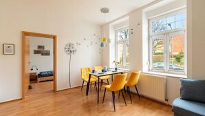 Ein helles Zimmer mit Holzboden, einem Esstisch mit gelben Stühlen, einem grauen Sofa und einem Fenster mit Blick auf einen Garten.